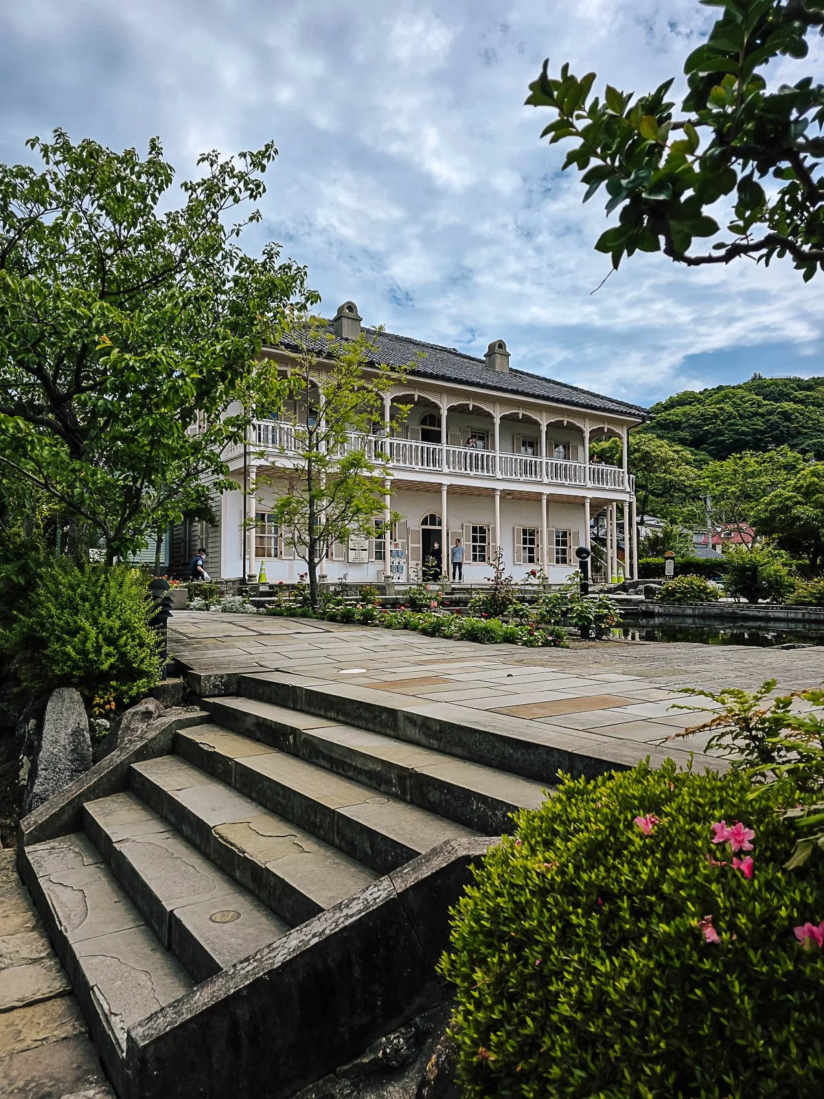 A colonial style house in Nagasaki Glover Garden. The house is white with a wrap around balcony on the first floor and surrounded by trees and gardens