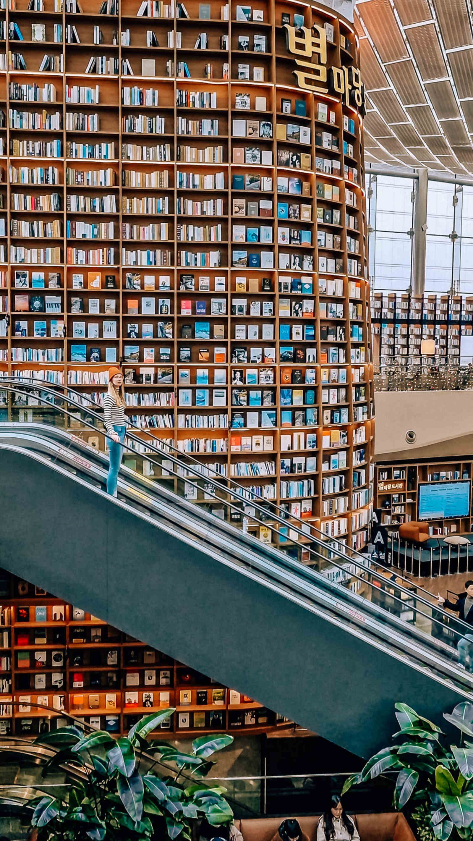 a huge atrium filled with floor to ceiling bookshelves with a woman going down an escalator in front of the shelves