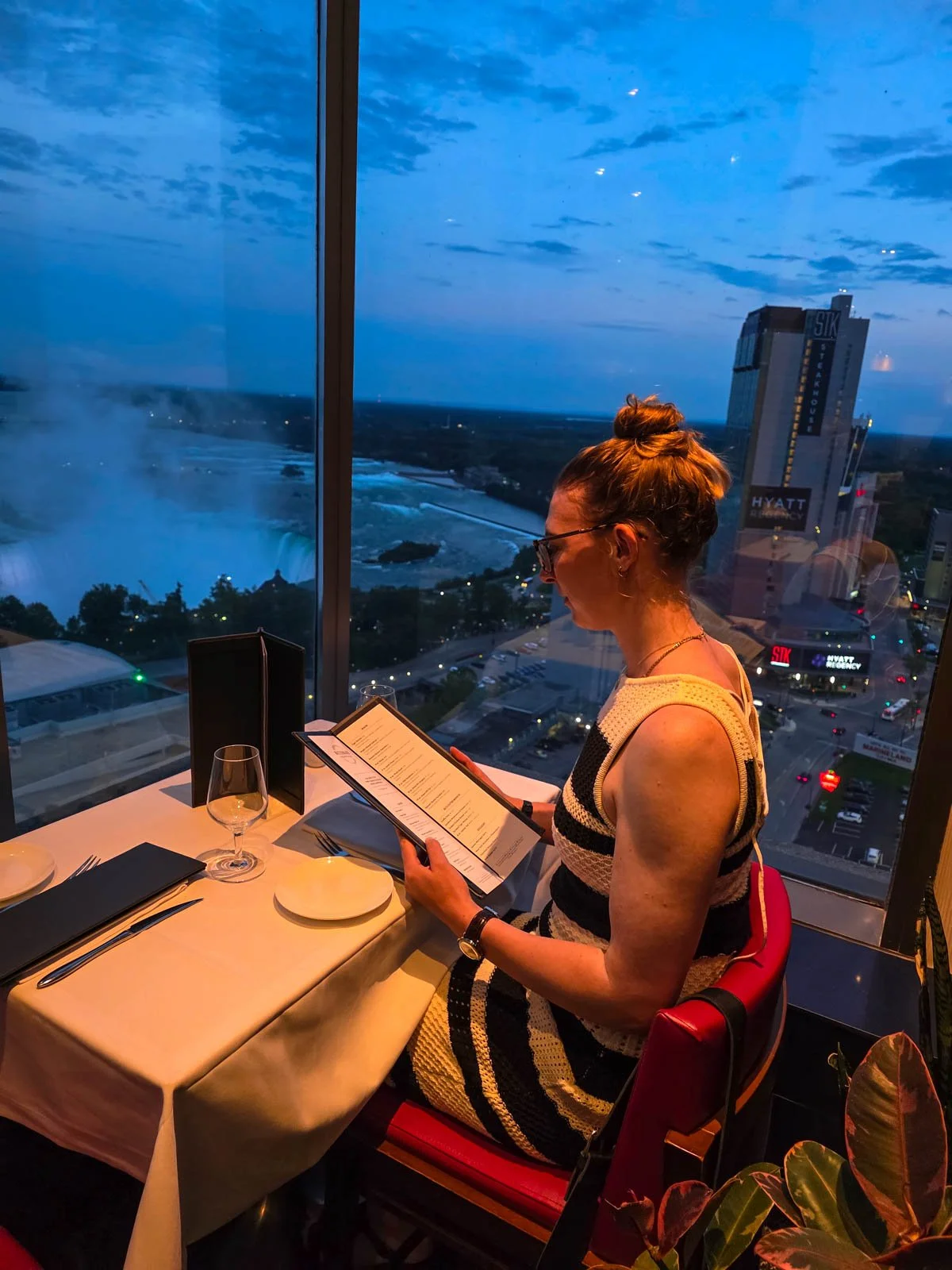 Helena in a black and white dress sitting in a restaurant reading menu with the window view of Niagara Falls in the background