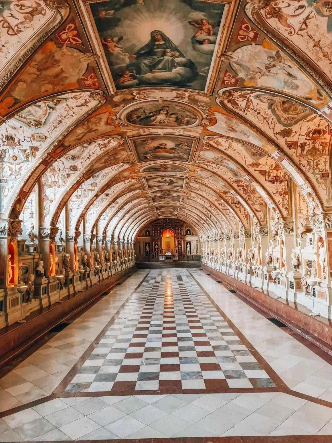 A large hallway with ornately painted ceilings and walls, decorated fixtures,  many statues lining the sides, and a tiled floor