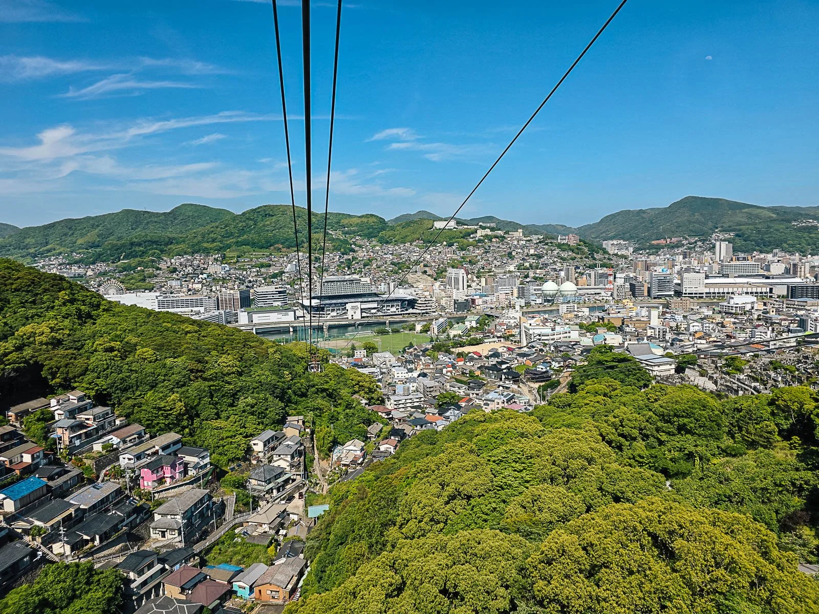 A view looking down on a hilly valley with a city and lots of hill covered trees. There are wires at the top of the photo as it's taken from an ascending cable car