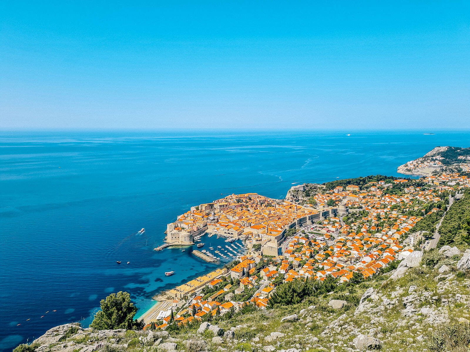 View from a mountain on the coast looking down on Dubrovnik town with orange roofs and a stone city wall around the buildings. The sea and sky are vibrant blue