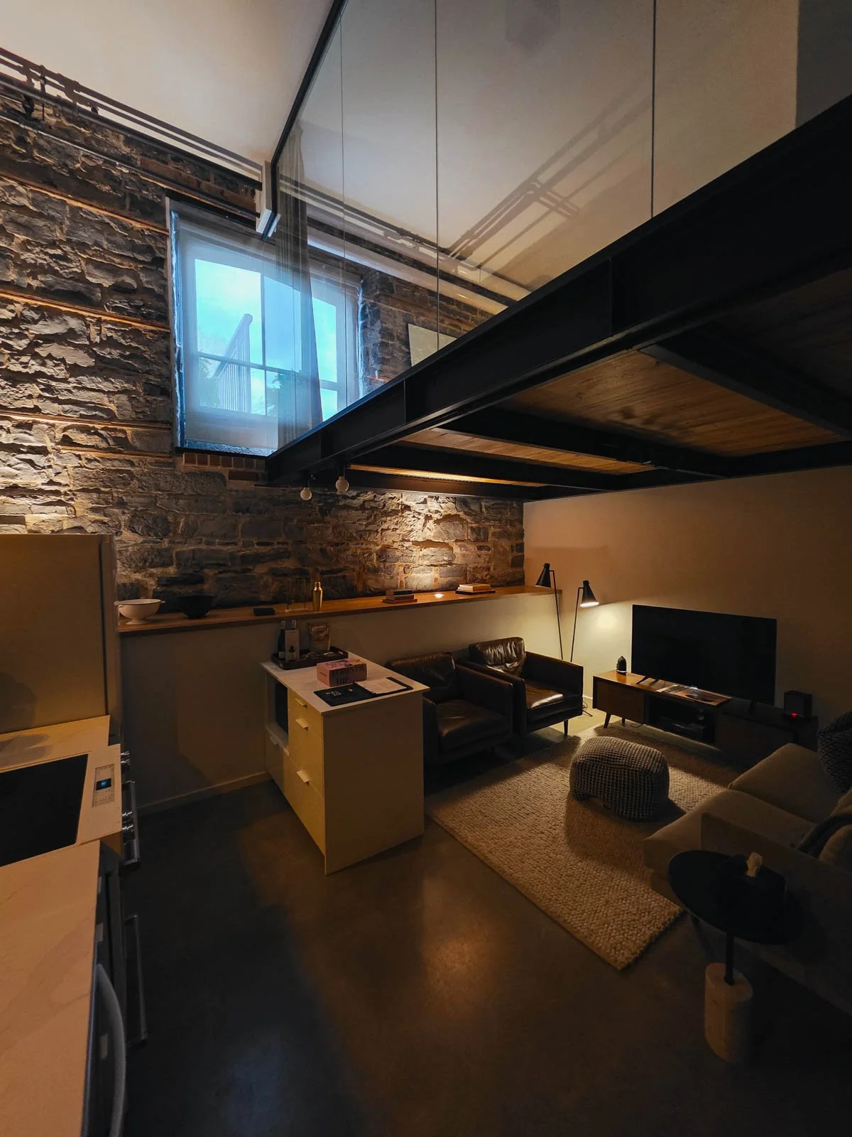 The interior of a large hotel room in a loft style with a kitchen, seating area with tv, and a large stone wall and a glass wall above where the loft level is