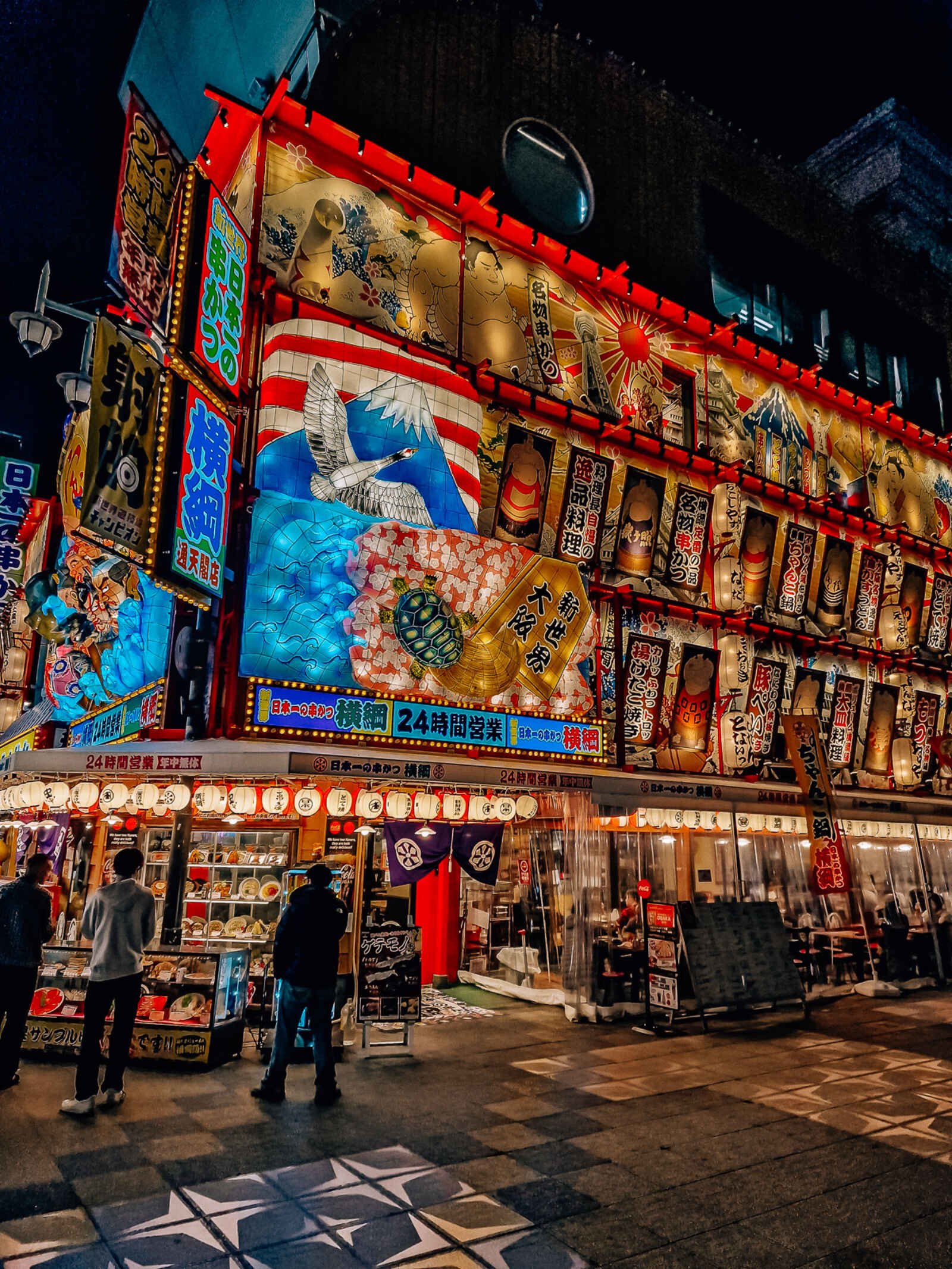At night time, a building covered in rectangular Japanese signs and lit up with a big picture of mount fuji and an eagle. Small lanterns line the entrance with people milling around in front