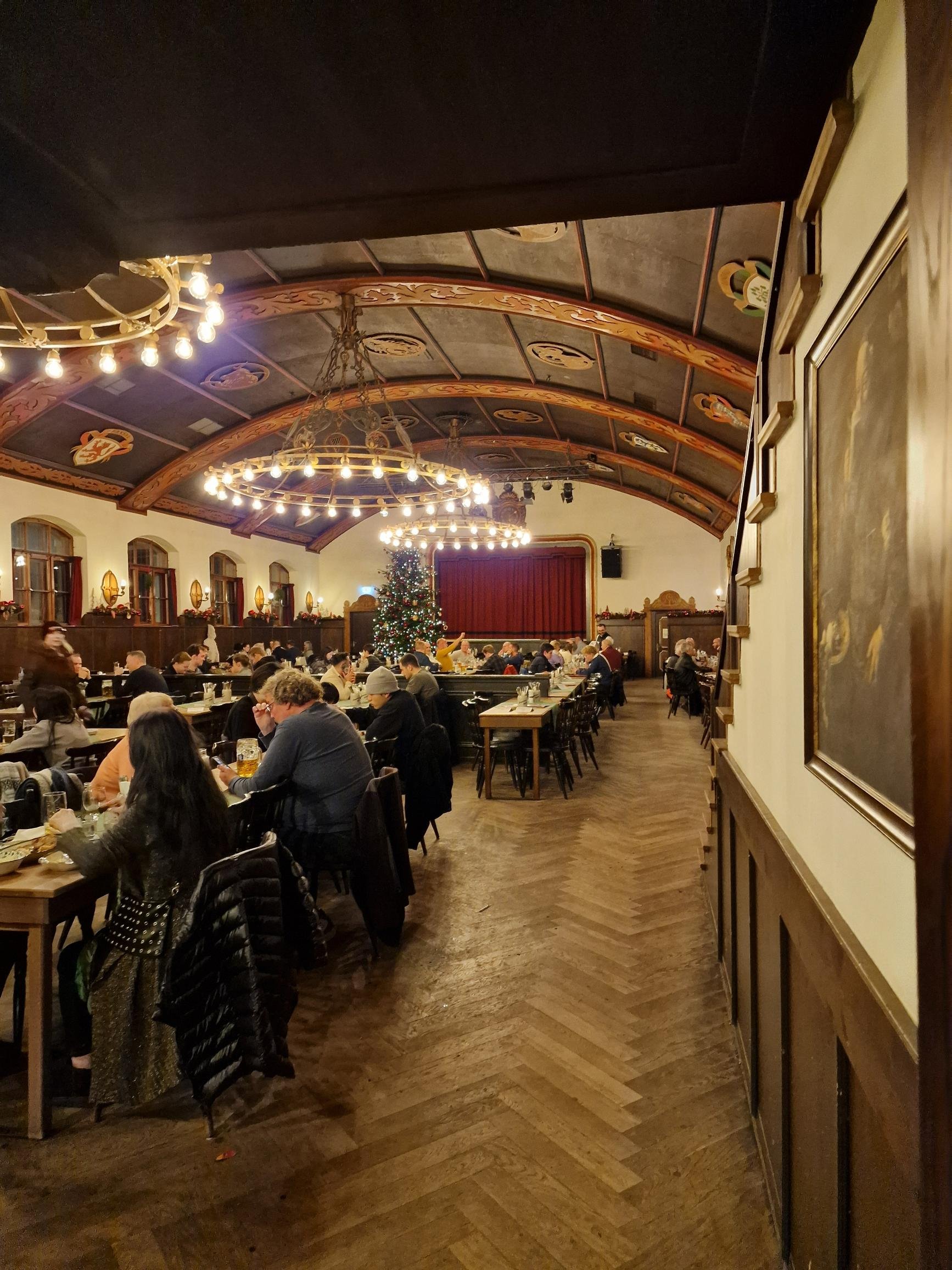 A large dark wood beer hall with many tables, large light fixtures, and red curtained stage at the far end