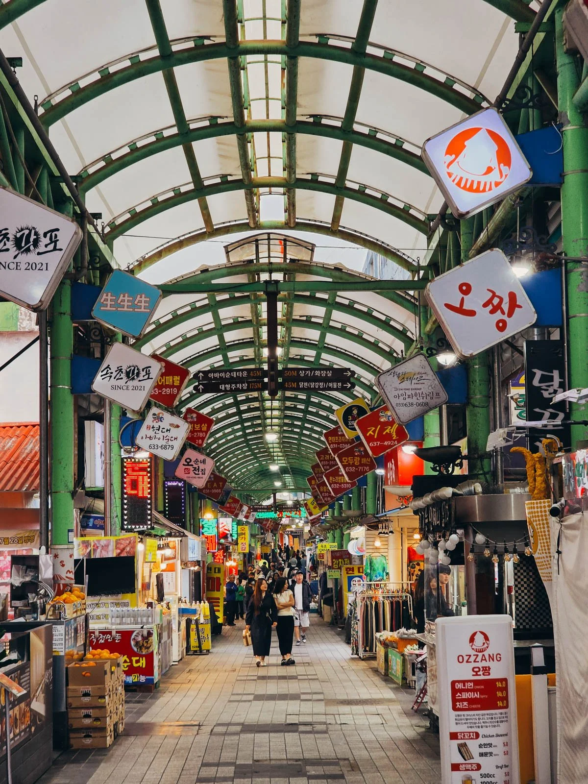 An indoor market row with green pillars and roof. Many food stall with colourful signs and food stands line either side of the alley