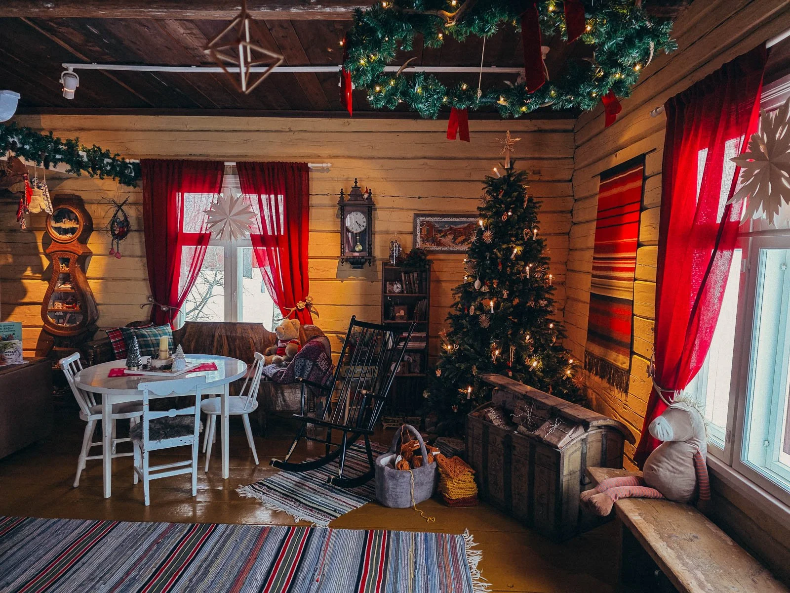 A large wooden cabin room decorated with a Christmas tree, red curtains, a wooden chest, and many other Christmas decorations