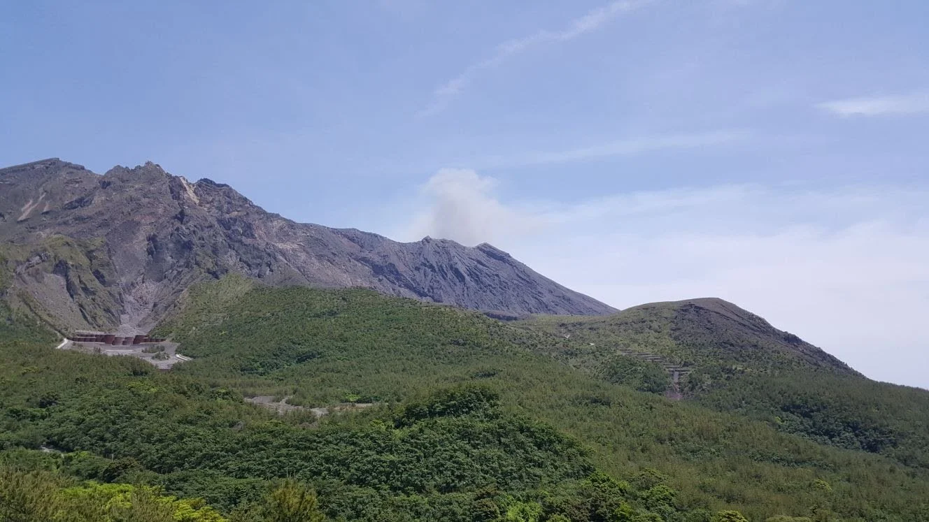 Looking up a large green and rocky hill to the top where a small plume of smoke is visible from the top of the volcano