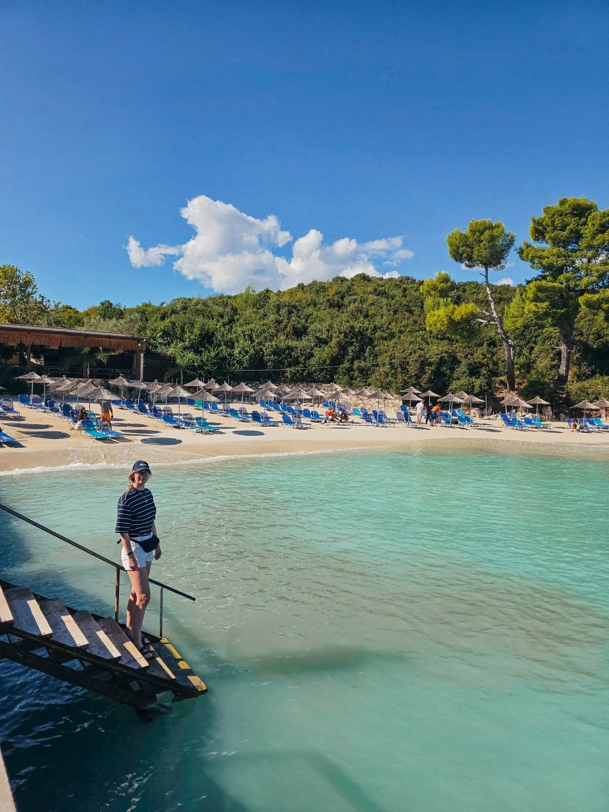 Helena standing on steps which lead into clear turquoise water. A white sanded beach with many beach chairs and umbrellas can be seen in the distance