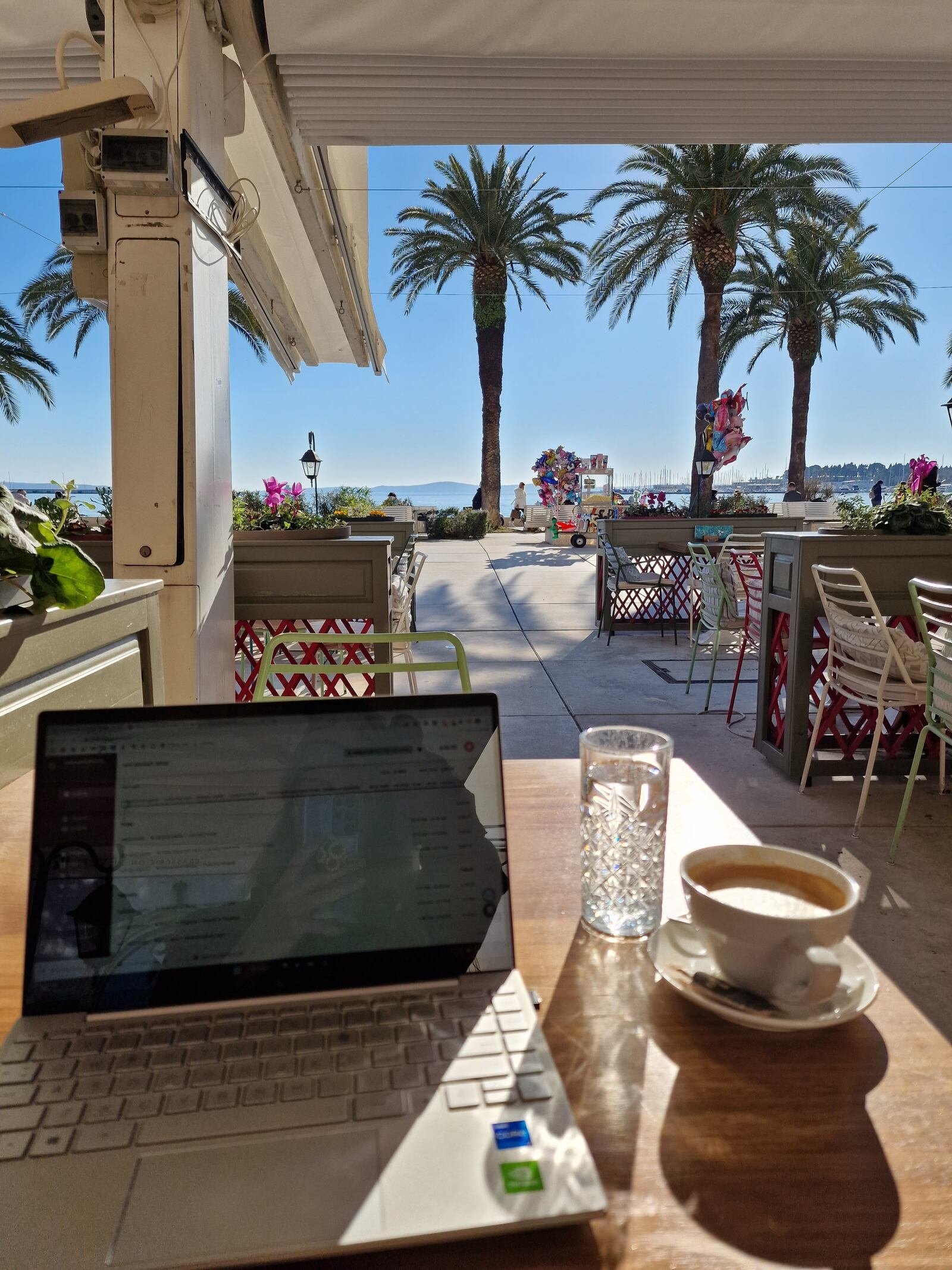 a laptop on a cafe table with coffee and water next to it, the view is outside of a sea harbour with blue sky and palm trees