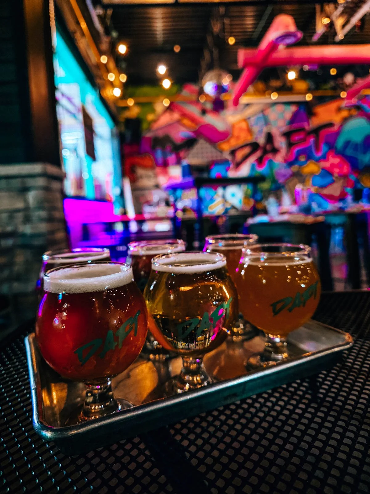 A silver tray on a table with 6 small round glasses of beer in a tasting flight, the room behind is neon and dark with graffiti art on the wall at a brewery