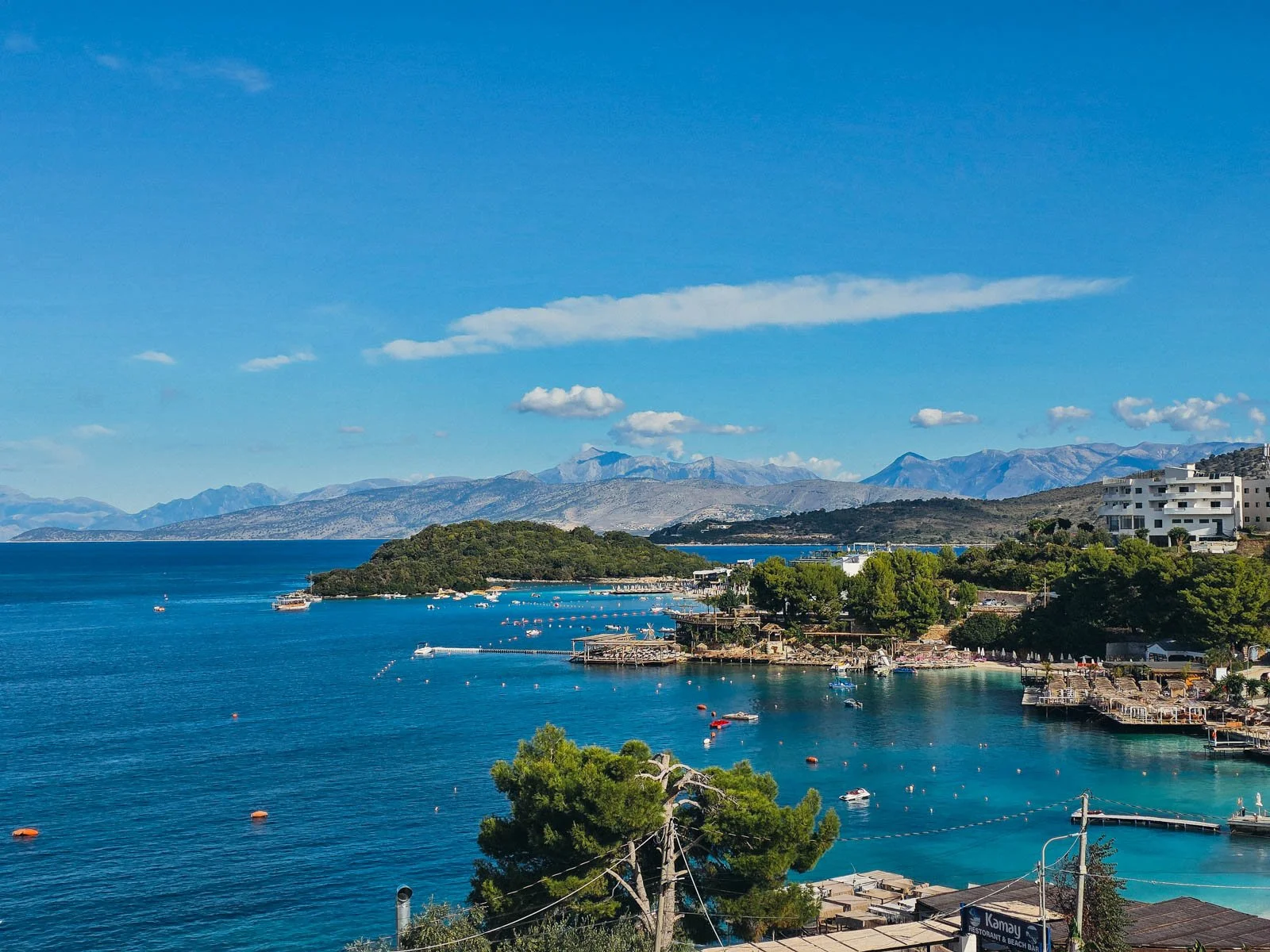 The coast of  a seaside town with many docks along clear blue water. There's a small tree coved island and mountains in the distance