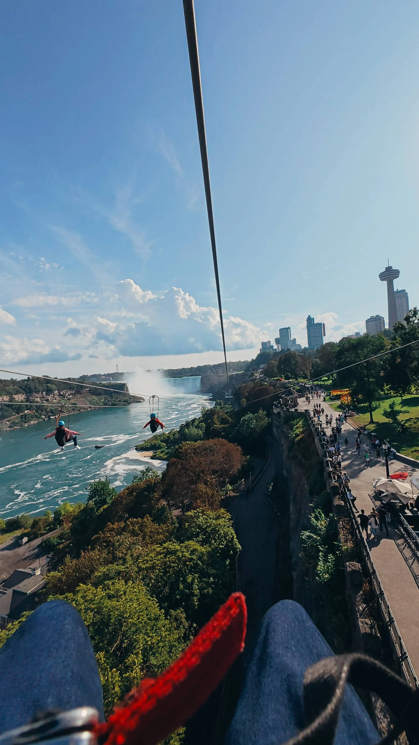 A perspective from someone ziplining over some trees next to a river with a view of Niagara Falls in the distance and three other zipliners either side
