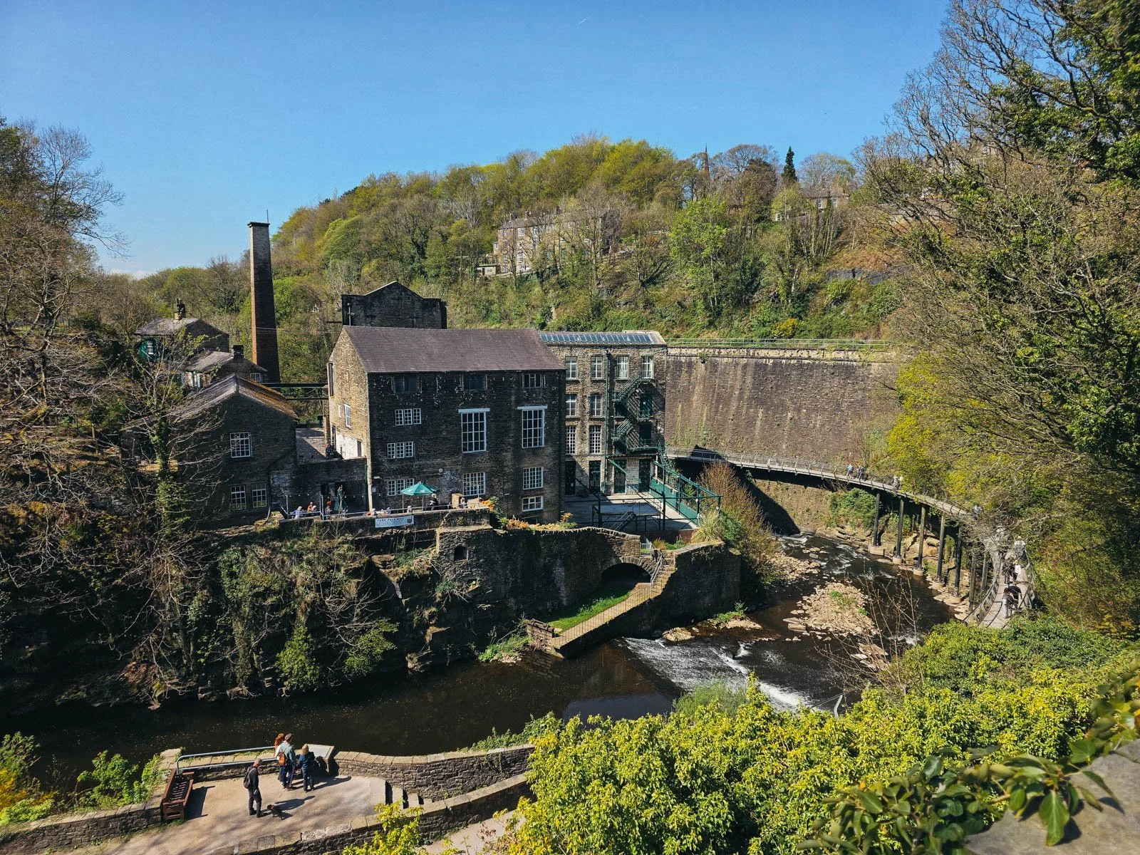 Looking down on a riverside park in New Mills with a gorge with a suspended walkway curving along the wall on the right, in the middle is a large, old converted mill, in the town of New Mills in High Peak - a day trip from Manchester