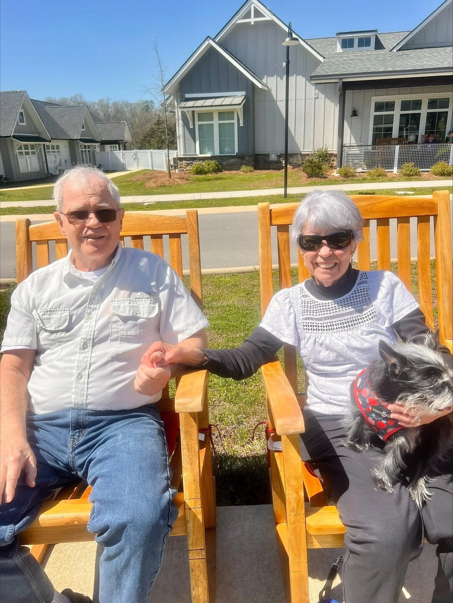 An elderly couple enjoys a sunny day outside on rocking chairs.