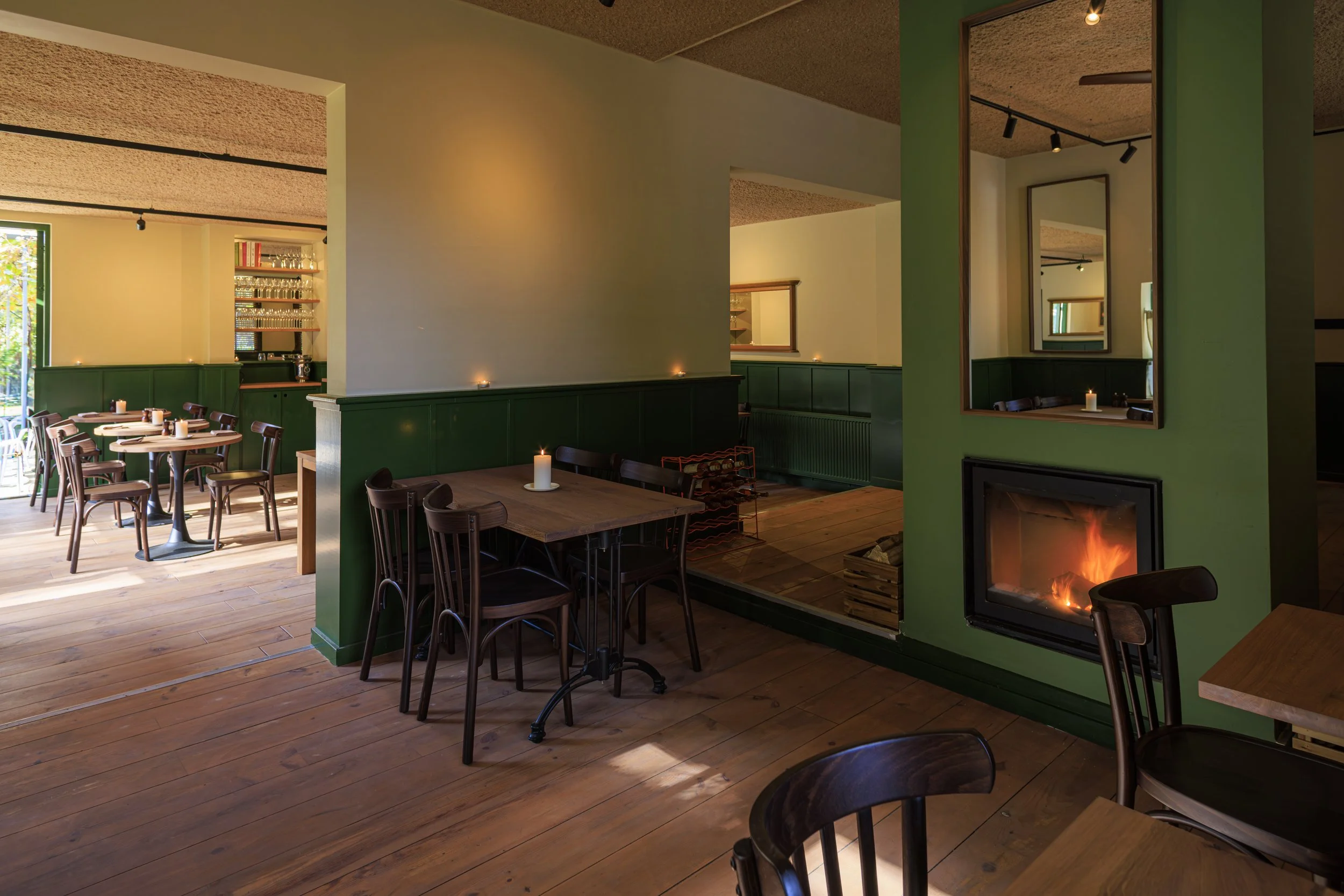 Interieur van een gezellige restaurant met houten vloeren, groene en witte muren, kaarslicht, een haardvuur, en houten tafels en stoelen.