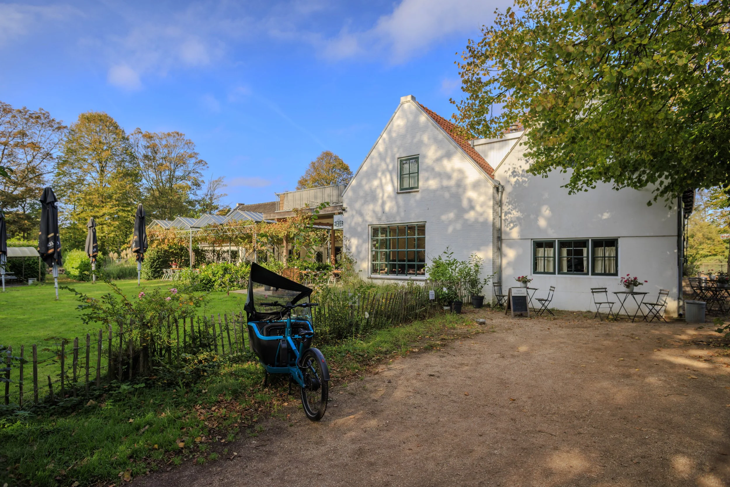 Een wit gebouw met een rode dak, omgeven door een tuin met groene planten, meerdere tuinmeubelen en parasols, en een bakfiets voorop de tuin. De lucht is blauw met wat wolken.