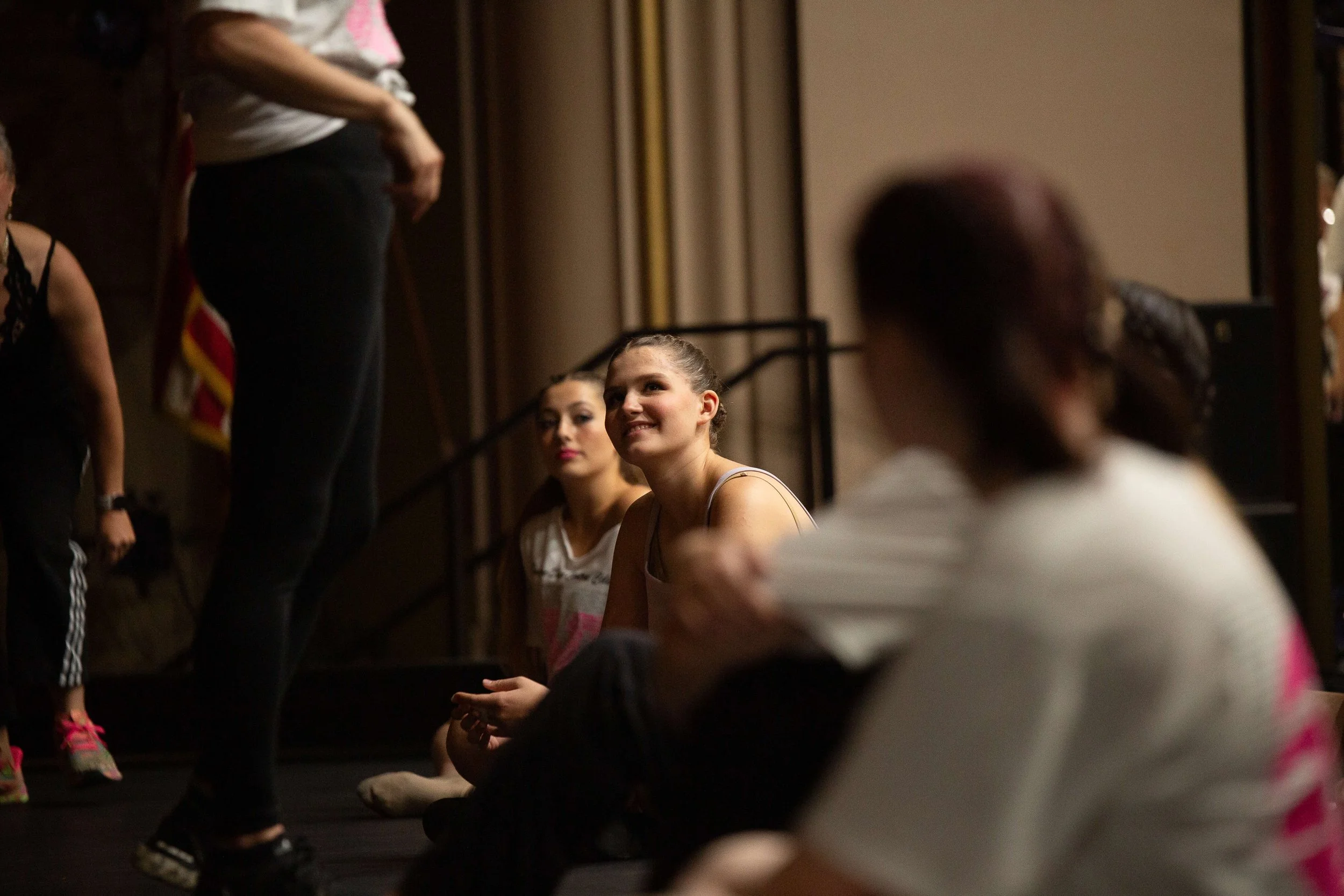 A group of young girls sitting on the floor, listening attentively, with a woman standing nearby in a dark room.
