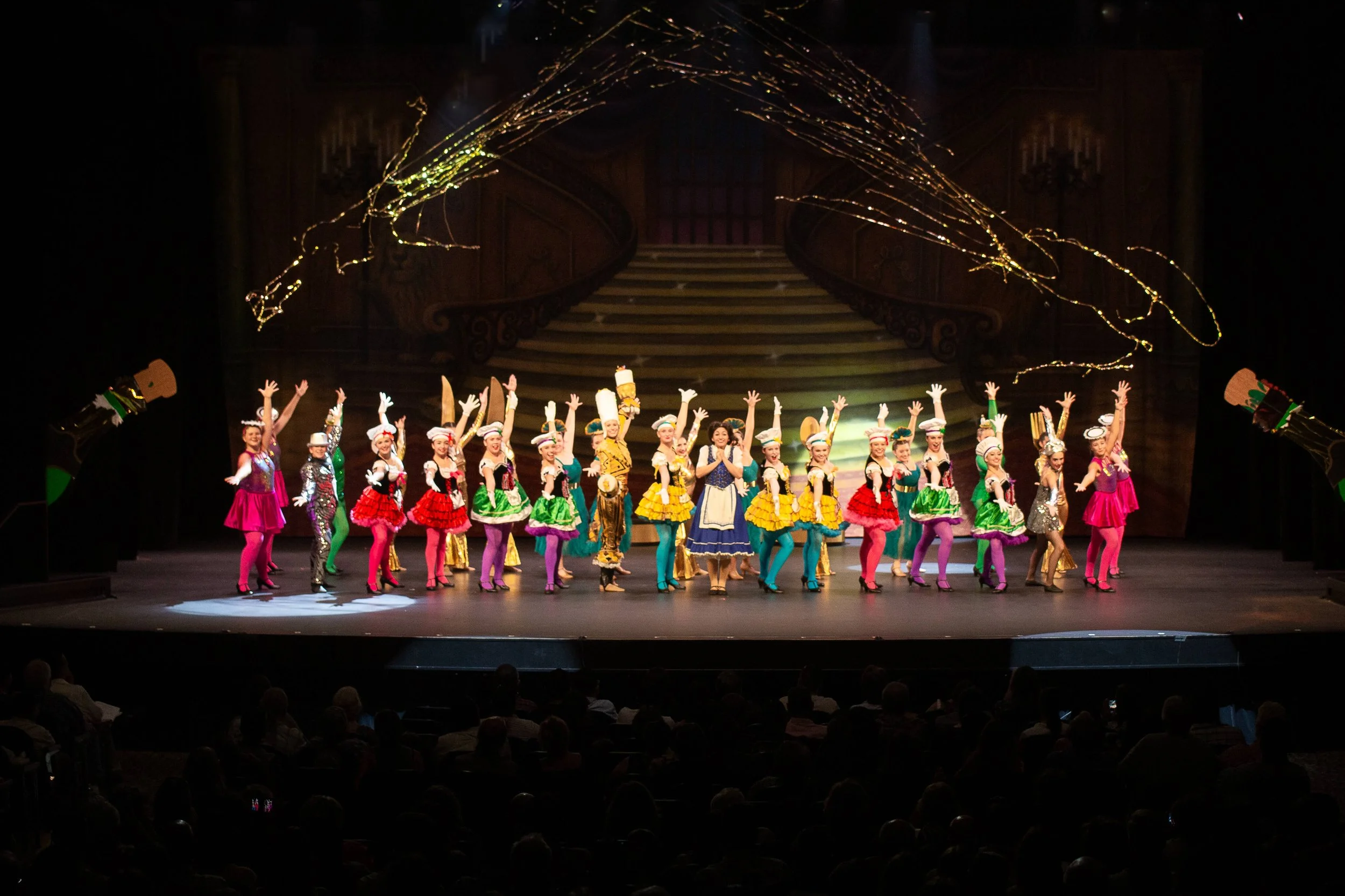 A group of performers dressed in colorful costumes are dancing on stage during a theatrical show with a grand staircase and ornate decor in the background, and golden light effects above them.