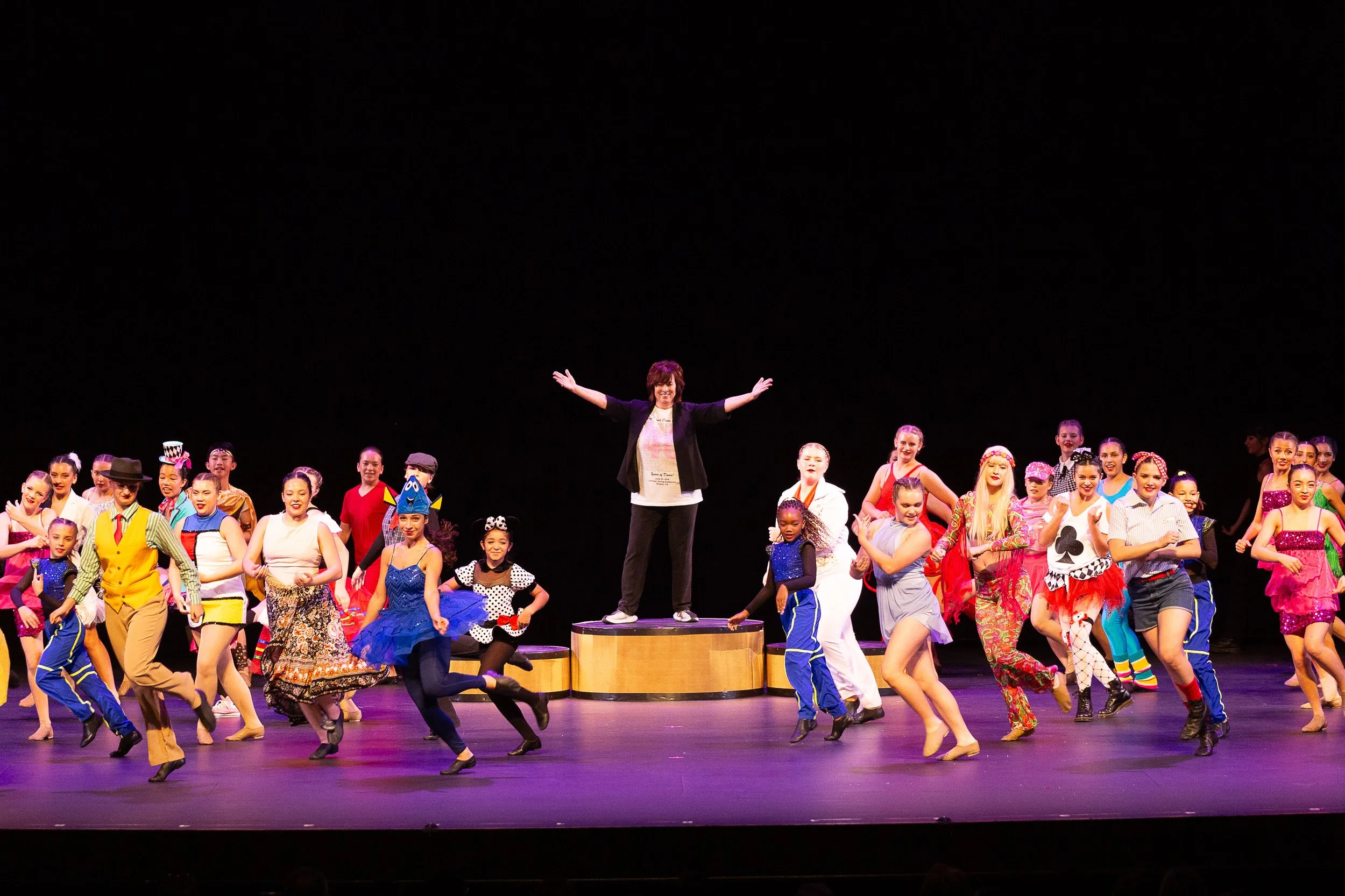 Group of young performers on stage, with their director in the center, dressed in colorful costumes, during a dance or musical theater performance.