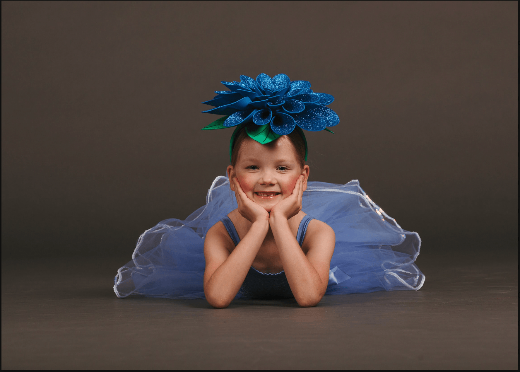 A young girl lying on the floor, propping her head up with her hands, smiling at the camera. She is wearing a blue dress with a tulle skirt, and a large blue flower headband with glitter. The background is dark gray.