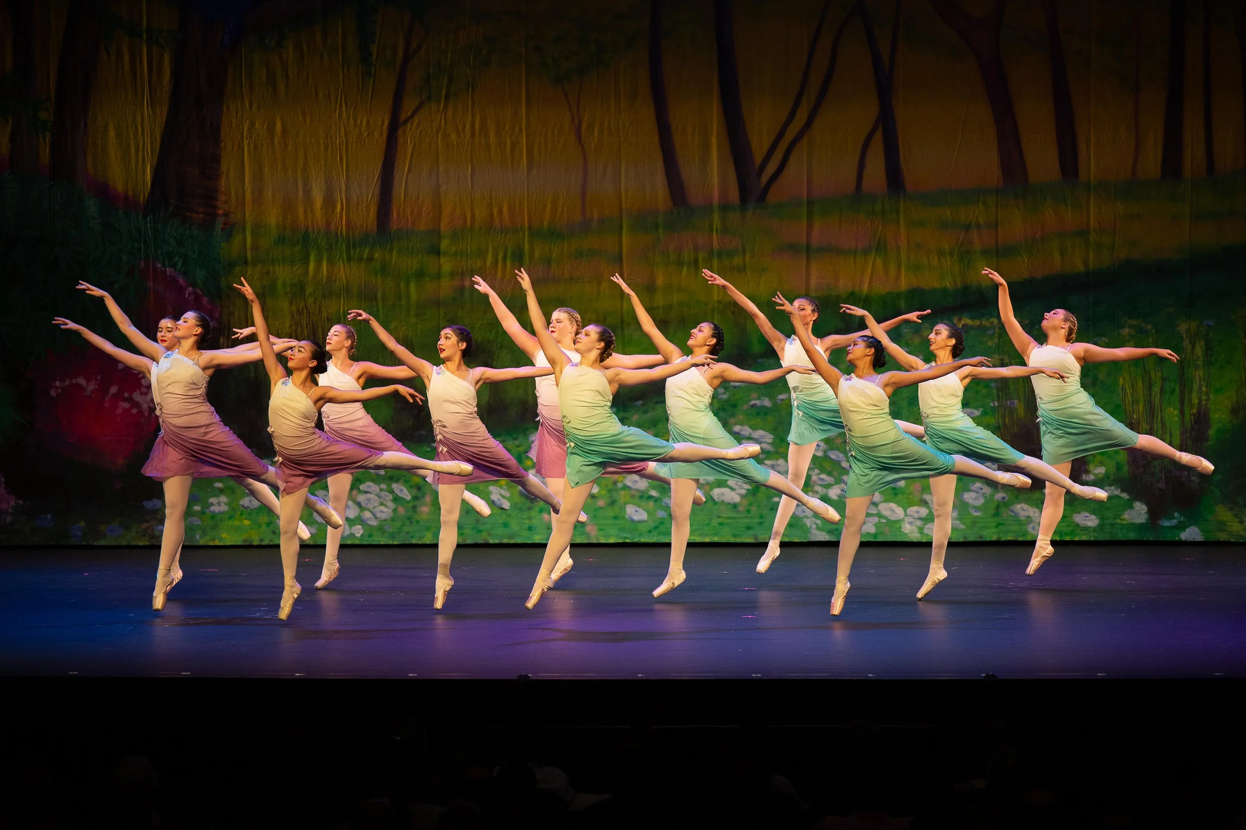 A group of ballet dancers in pastel costumes performing on stage against a scenic backdrop of trees and nature.