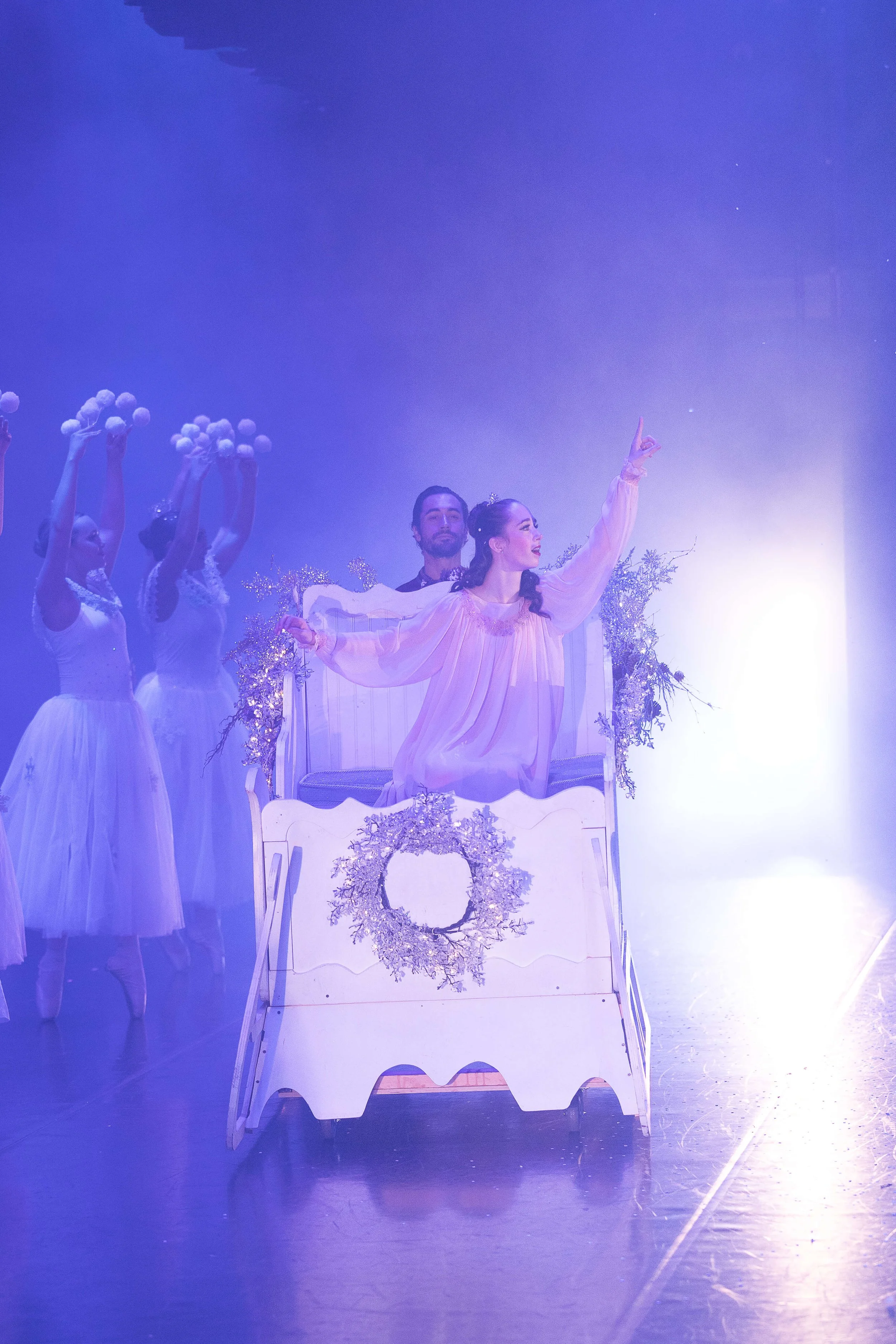 Stage scene with female ballet dancers in white tutus and a female singer or performer in a pink dress, sitting in a decorated bed with floral and wreath adornments, under purple lighting.