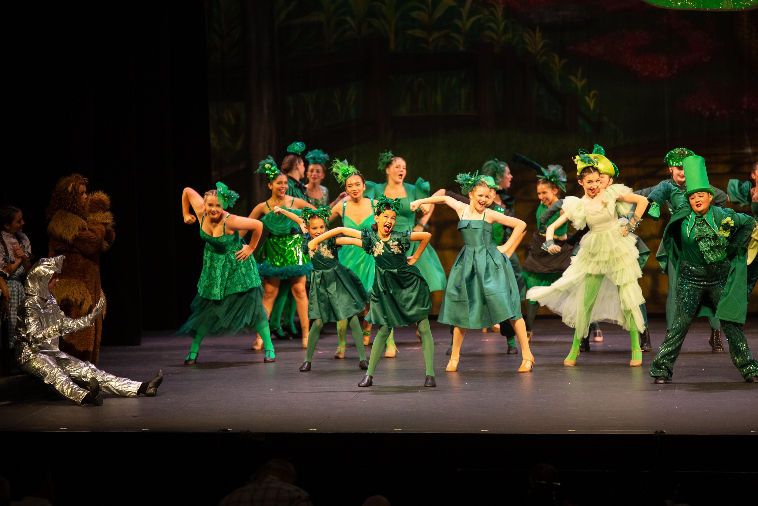 Children dressed in green costumes performing a dance on stage with a forest background, some characters in animal costumes are visible to the side.