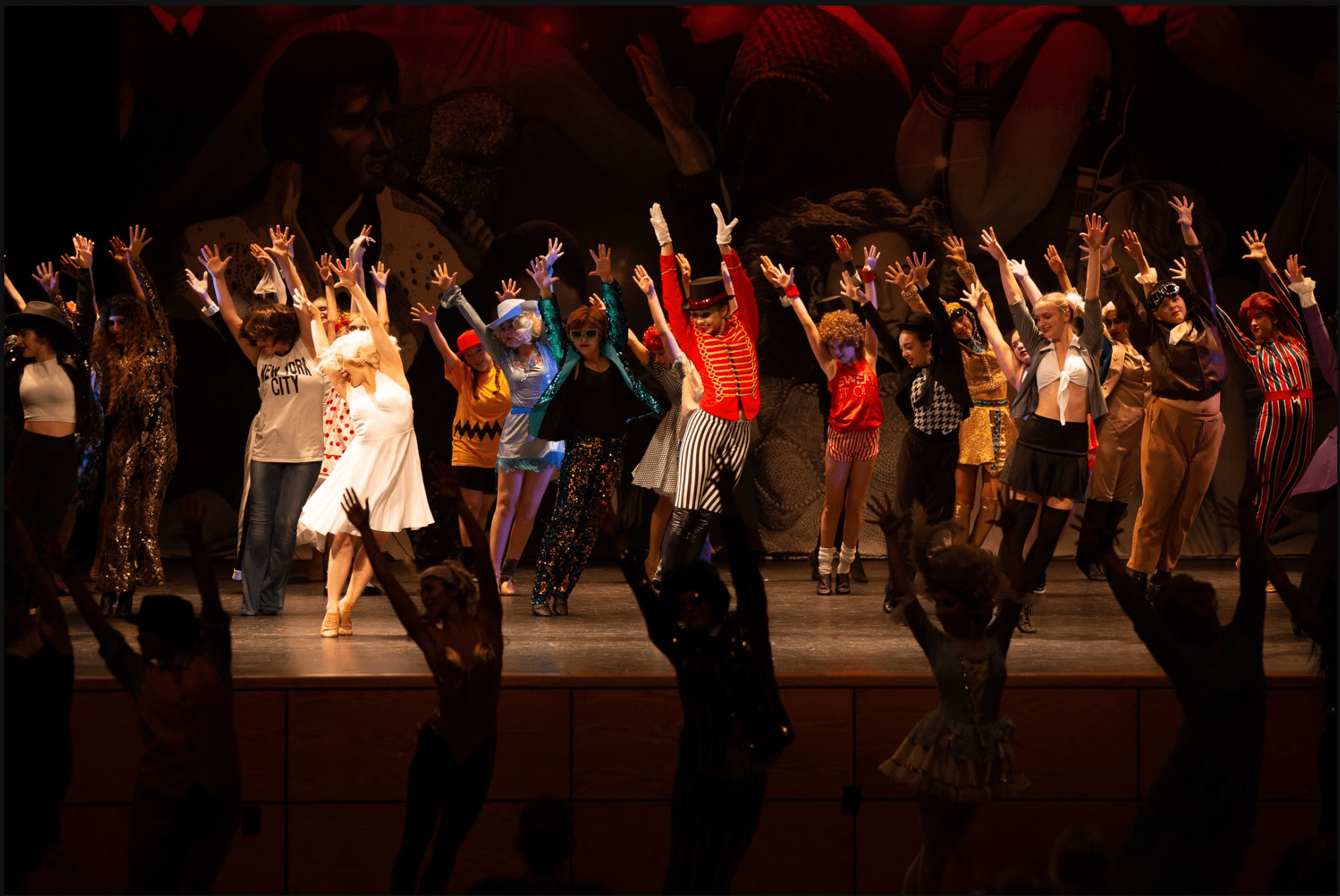Group of performers dressed in colorful costumes, dancing on stage with raised arms during a show.