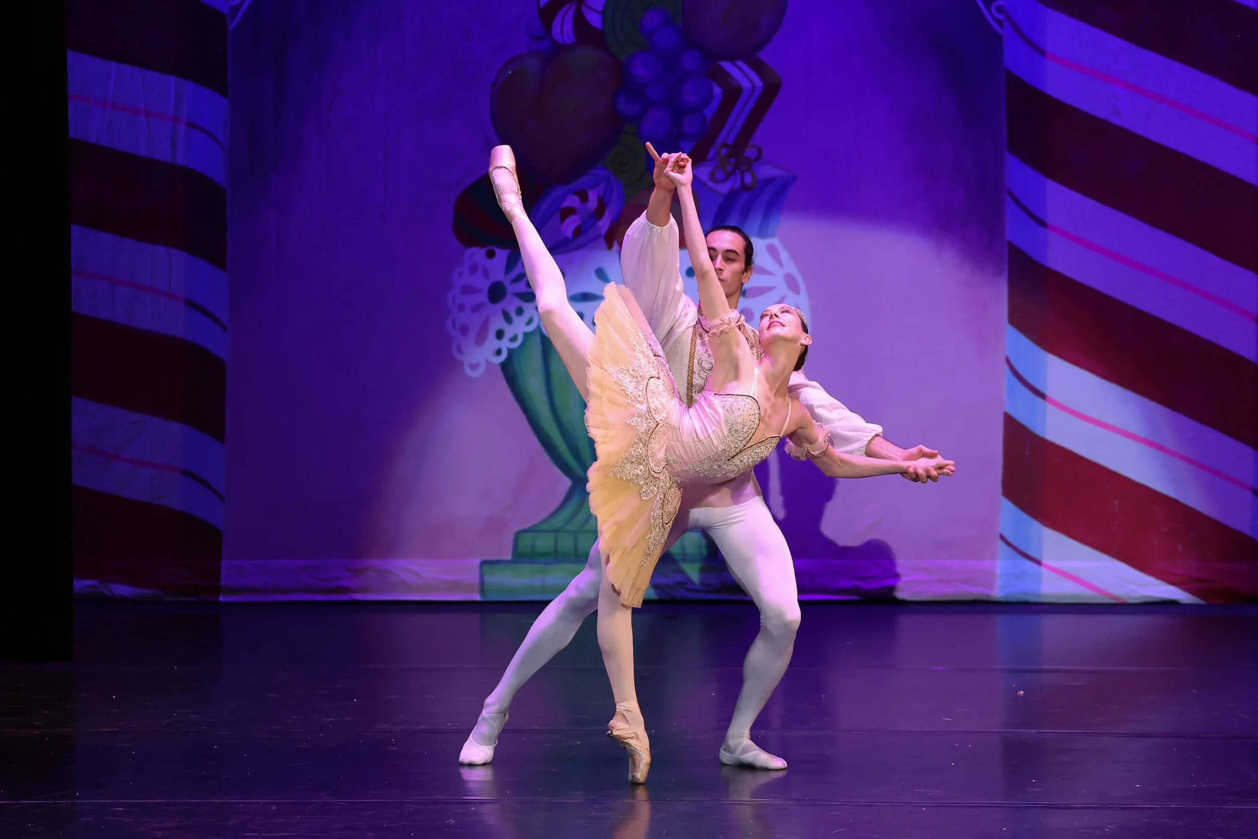 Two ballet dancers perform on stage, with the male dancer supporting the female dancer in an arabesque pose. The stage backdrop features colorful, abstract artwork.
