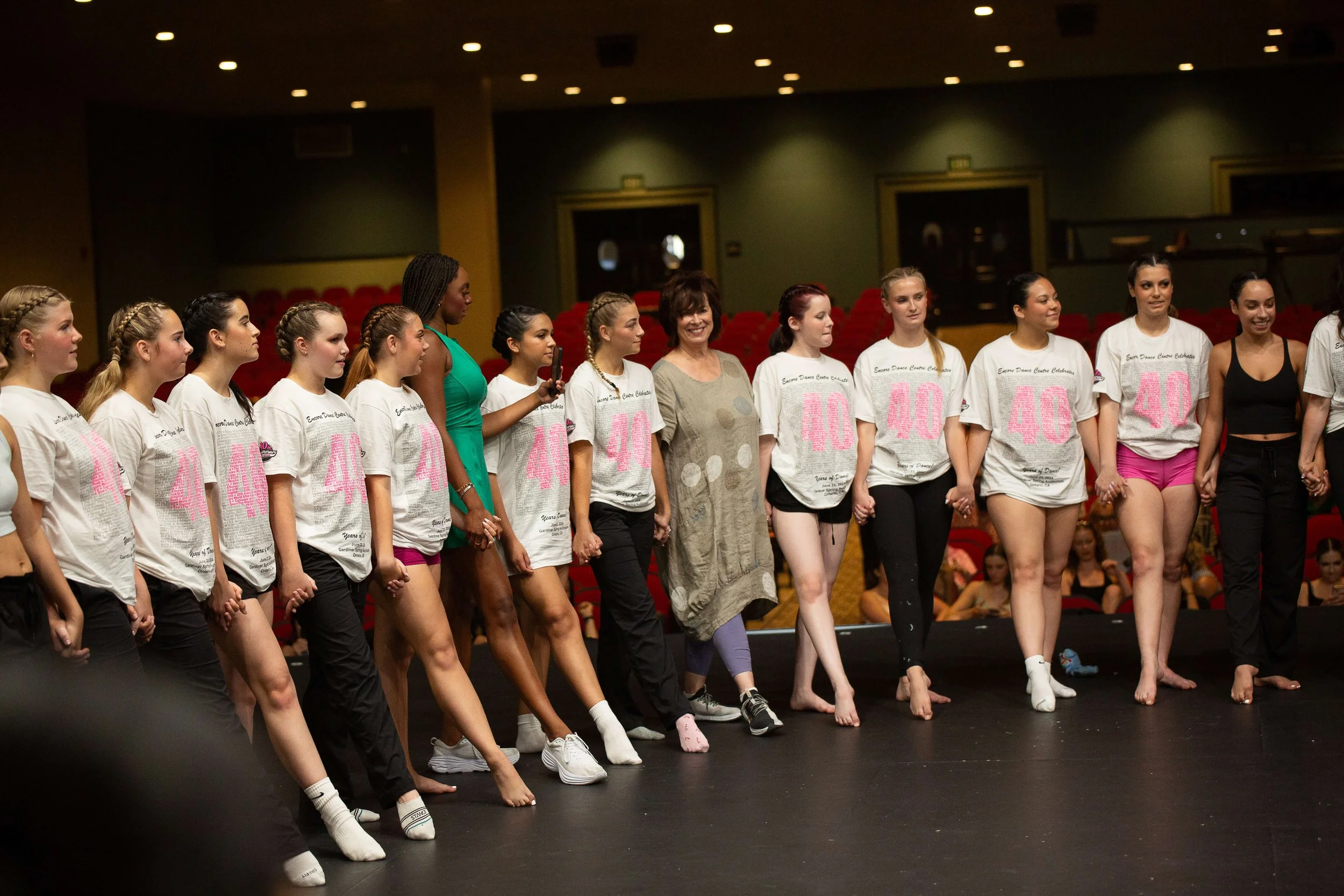 A group of female dancers and their instructor standing hand in hand on stage after a dance performance in a theater with red seats and green walls.