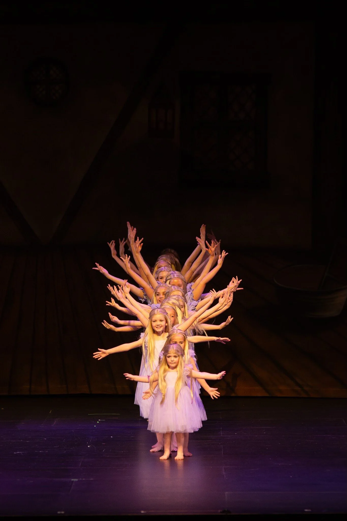 A group of young girls performing a dance on stage, arranged in a line with their arms extended out to the sides. They are wearing matching light-colored costumes with flowing skirts, and are positioned in a formation that creates the illusion of multiple arms extending from the front girl. The background is dark, emphasizing the performers.