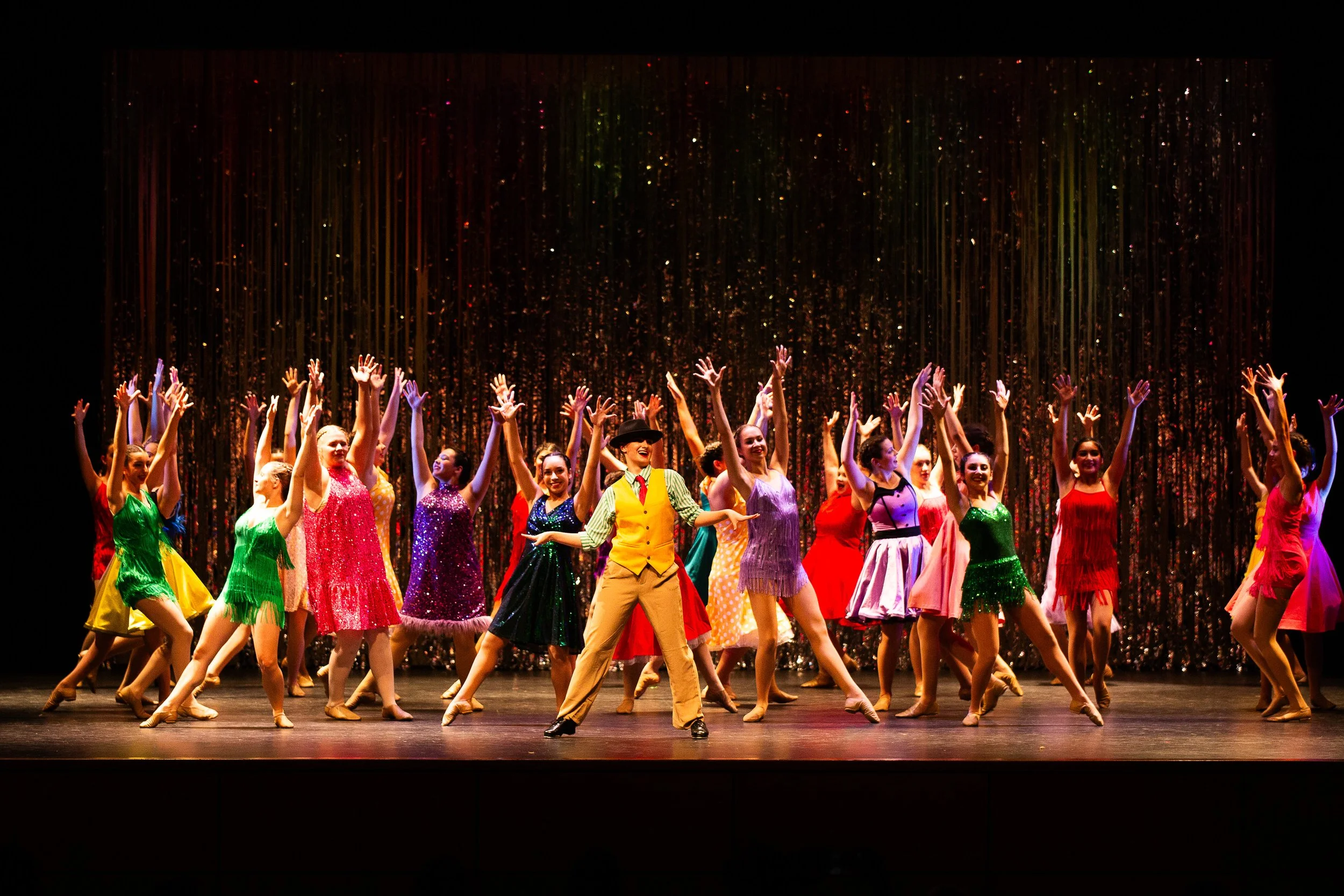 The image shows a group of performers on stage, dressed in colorful costumes, dancing with their arms raised, under a backdrop of shimmering curtain.