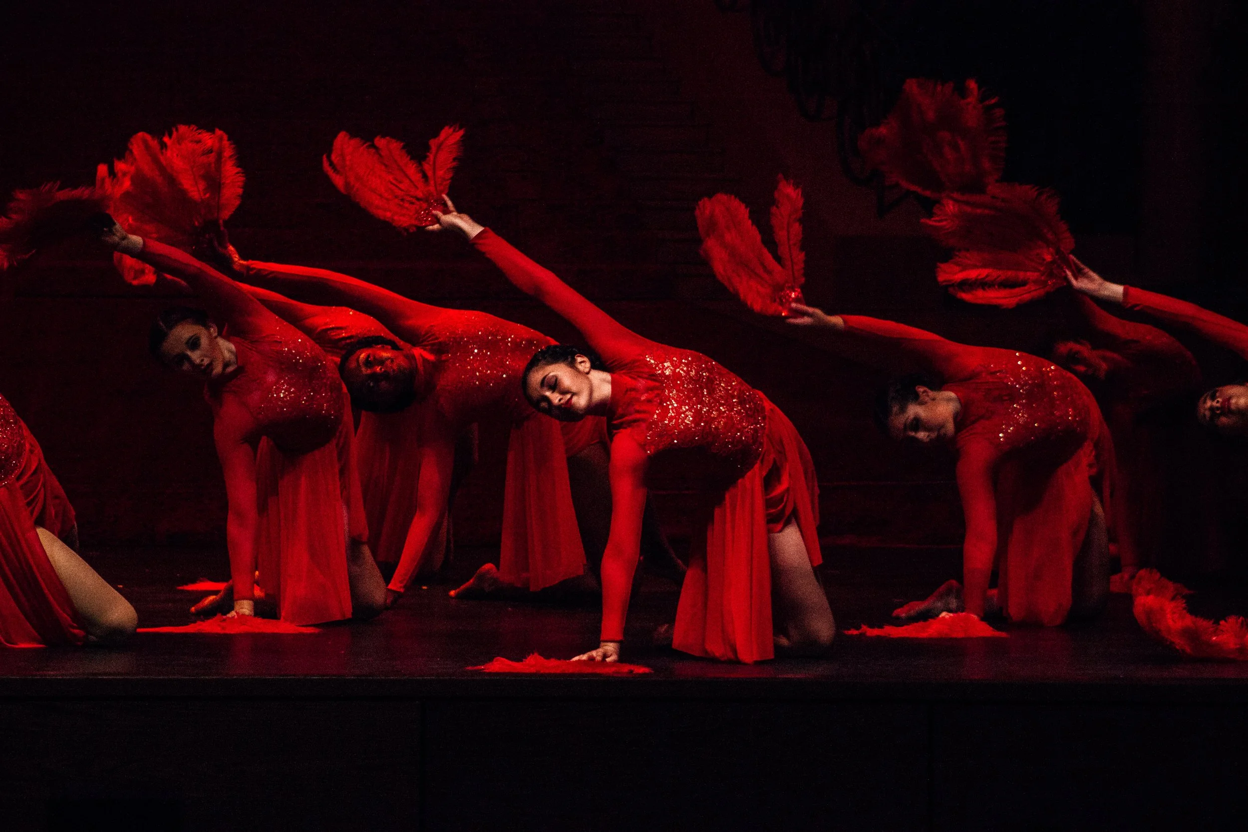 Performers dressed in red costumes, holding red fans, in a choreographed dance on stage with a dark background.