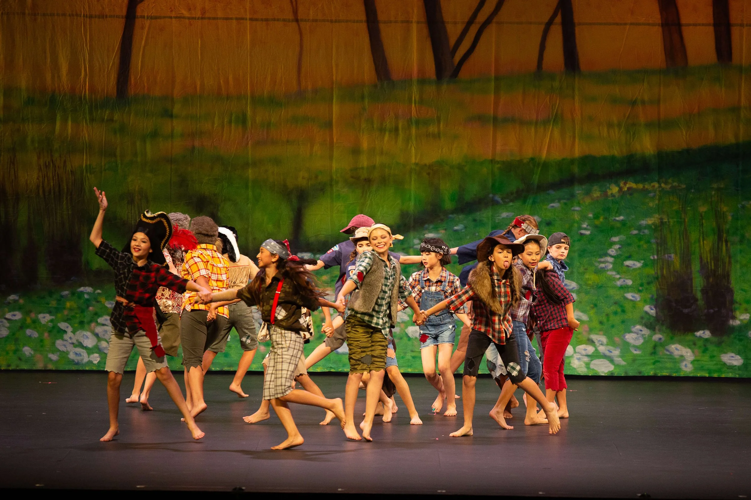 Children dressed in pirate and outdoor costumes perform a dance routine on stage with a forest backdrop.