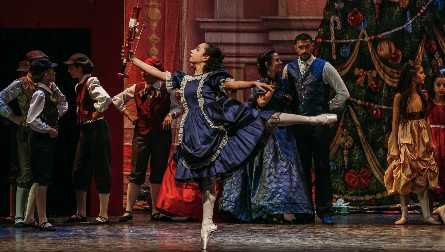 Ballet dancers performing on stage during a holiday-themed production with a decorated Christmas tree in the background.