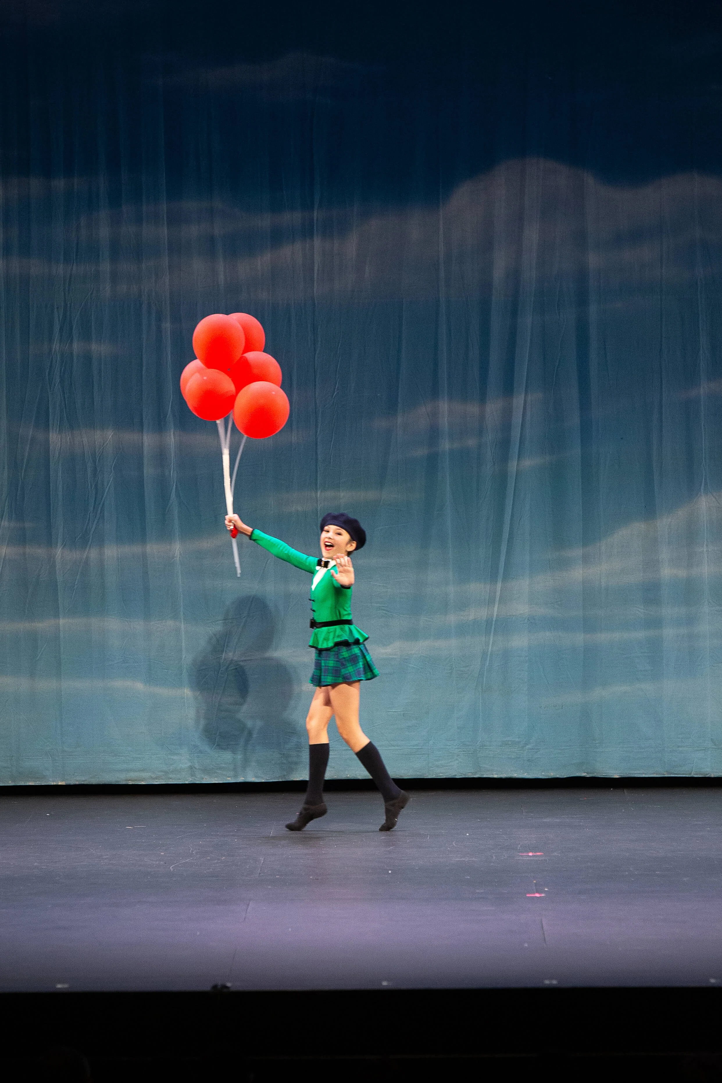 A girl dressed in a green top and plaid skirt, holding a bunch of red balloons, performing on stage with a dark curtain background.