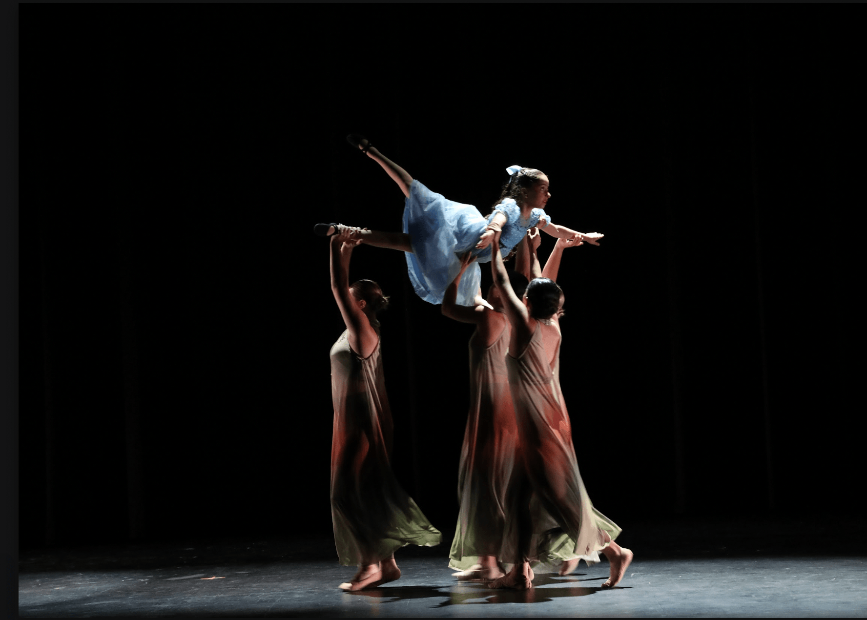 A group of five women performing a dance on stage with a dark background. One woman, dressed in a blue costume, is being lifted and extended horizontally by the others.