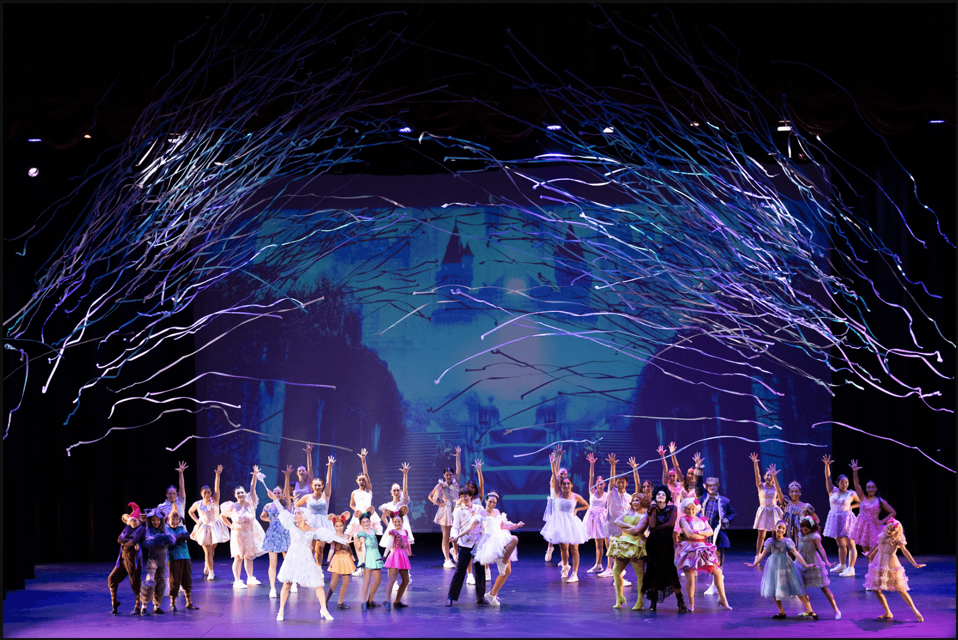 Large group of performers on stage dressed in colorful costumes, raising their hands in a synchronized dance, under a darkened theater with dramatic lighting and swirling light effects overhead, resembling a theatrical performance or ballet.