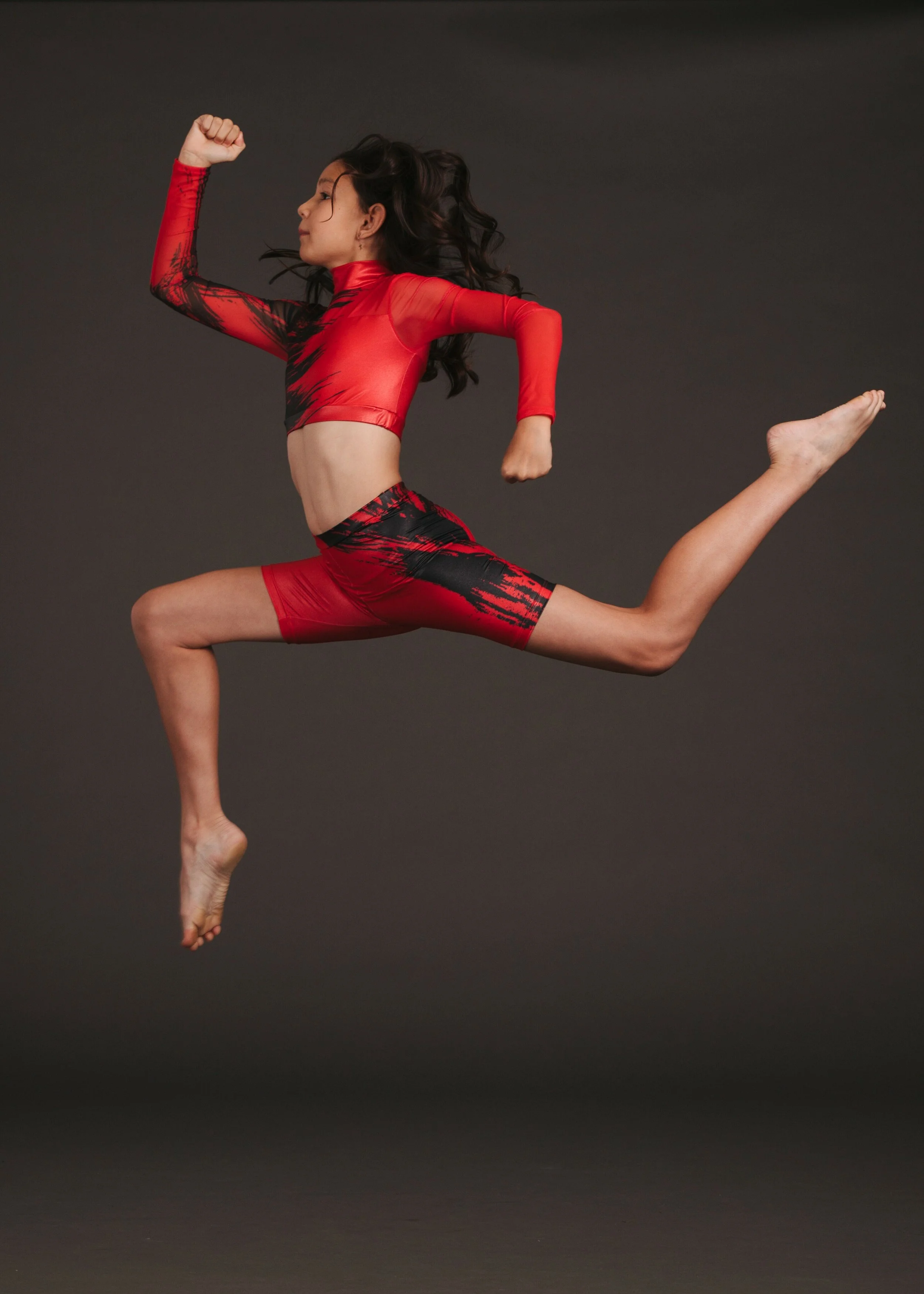 A woman in a red and black athletic outfit performs a leap, captured mid-air against a dark background.