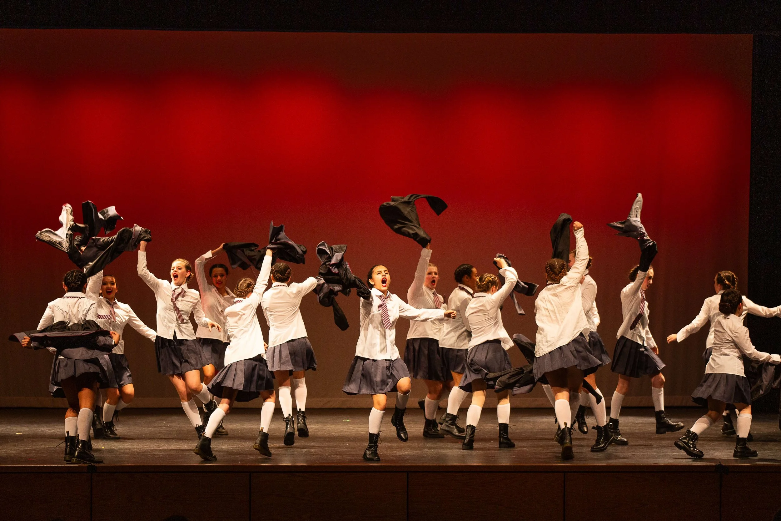A group of children is dancing on stage, waving black jackets in the air, wearing white shirts and dark skirts or pants, with a red background backdrop.