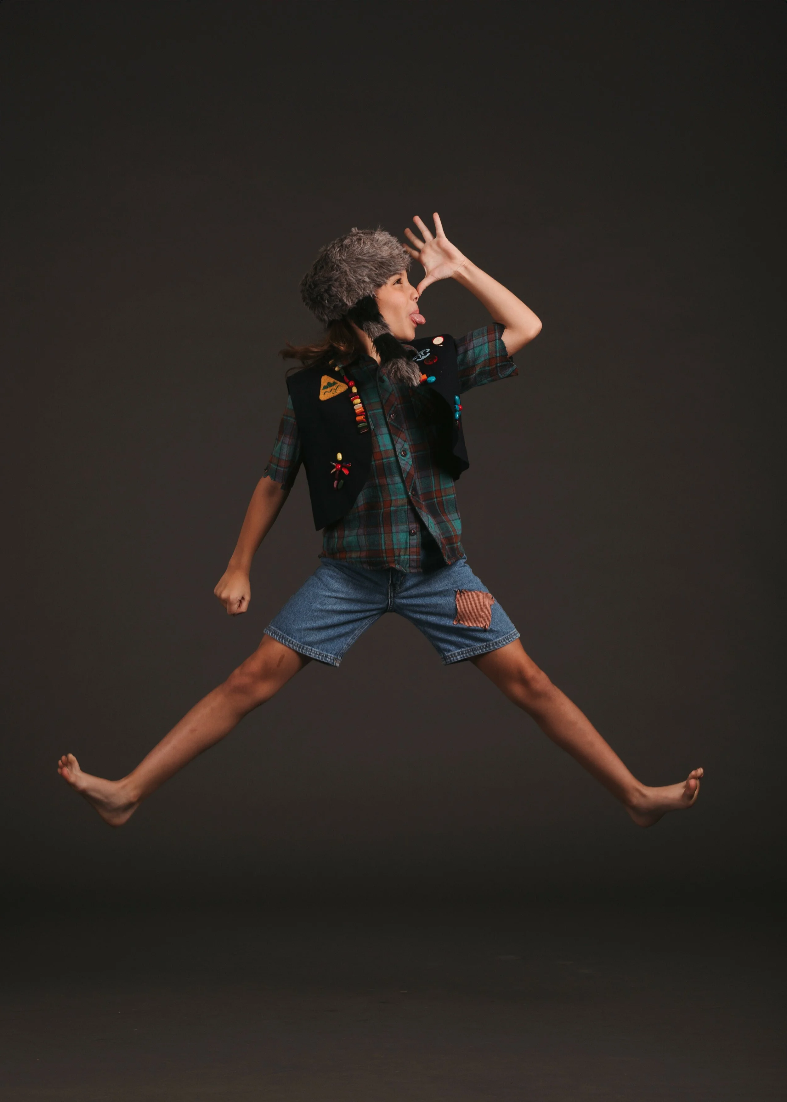 A woman wearing a plaid shirt, denim shorts, a furry hat, and a vest with patches and decorations, is posing with her legs and arms spread wide and sticking out her tongue against a plain dark background.
