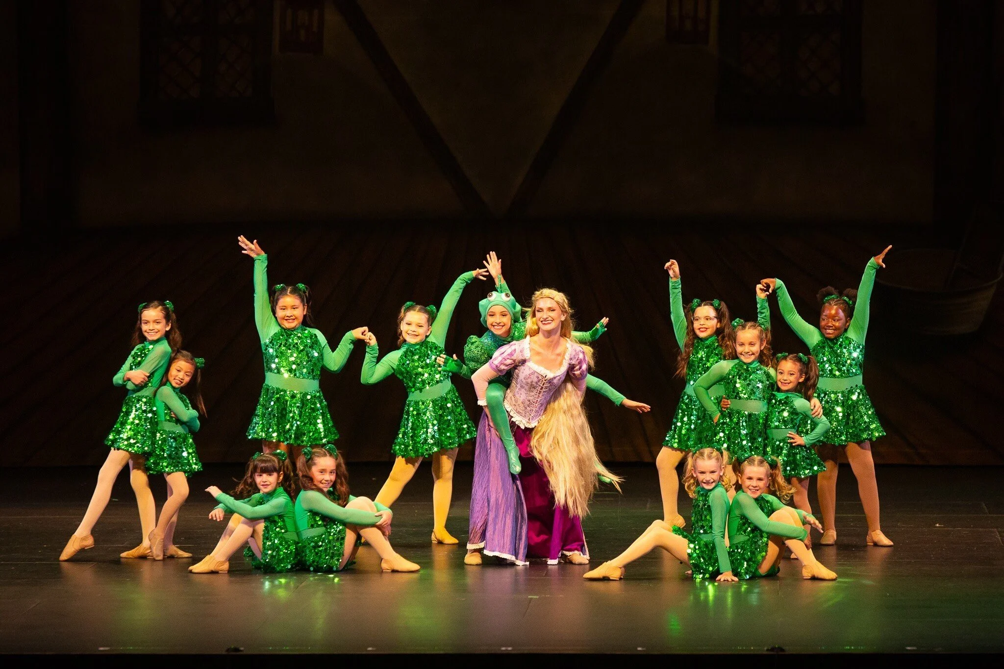 Group of young girls in green costumes and a woman dressed as Rapunzel on stage during a dance performance.