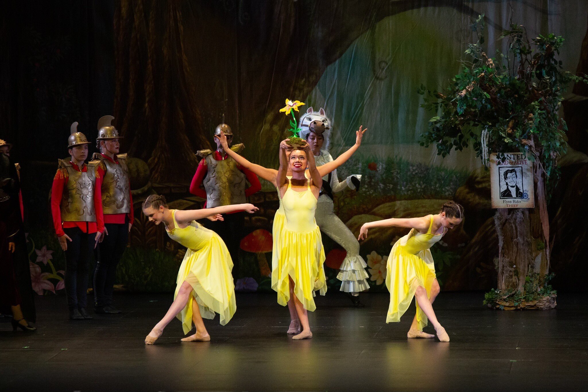 Ballet dancers on stage, some wearing yellow skirts and others in costumes resembling soldiers with helmets. One dancer in a yellow dress holds a flower-shaped prop on her head. The background features a dark forest scene.