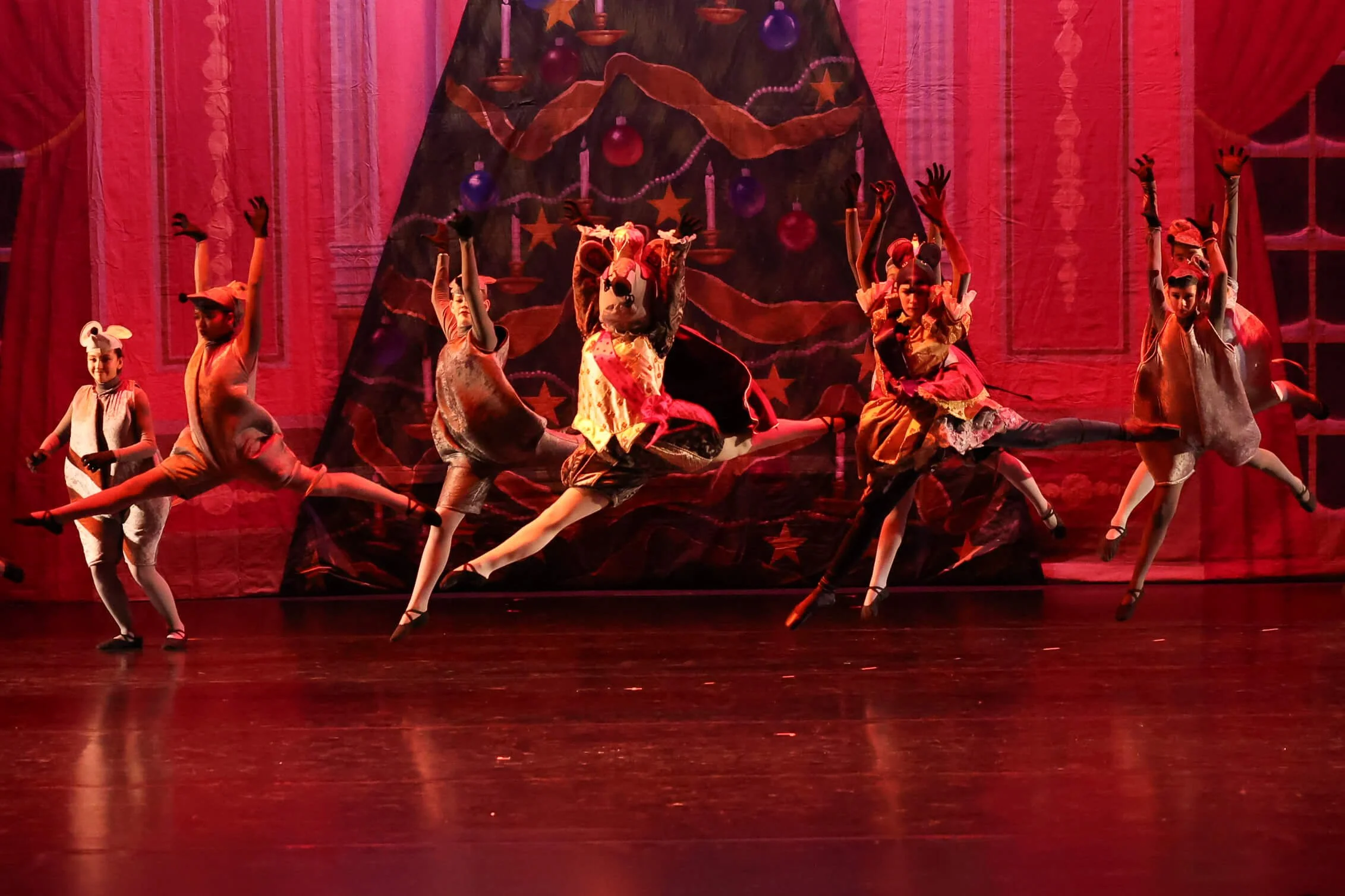 Children wearing costumes, some dressed as animals, performing a dance on stage with a red backdrop and Christmas-themed decorations.