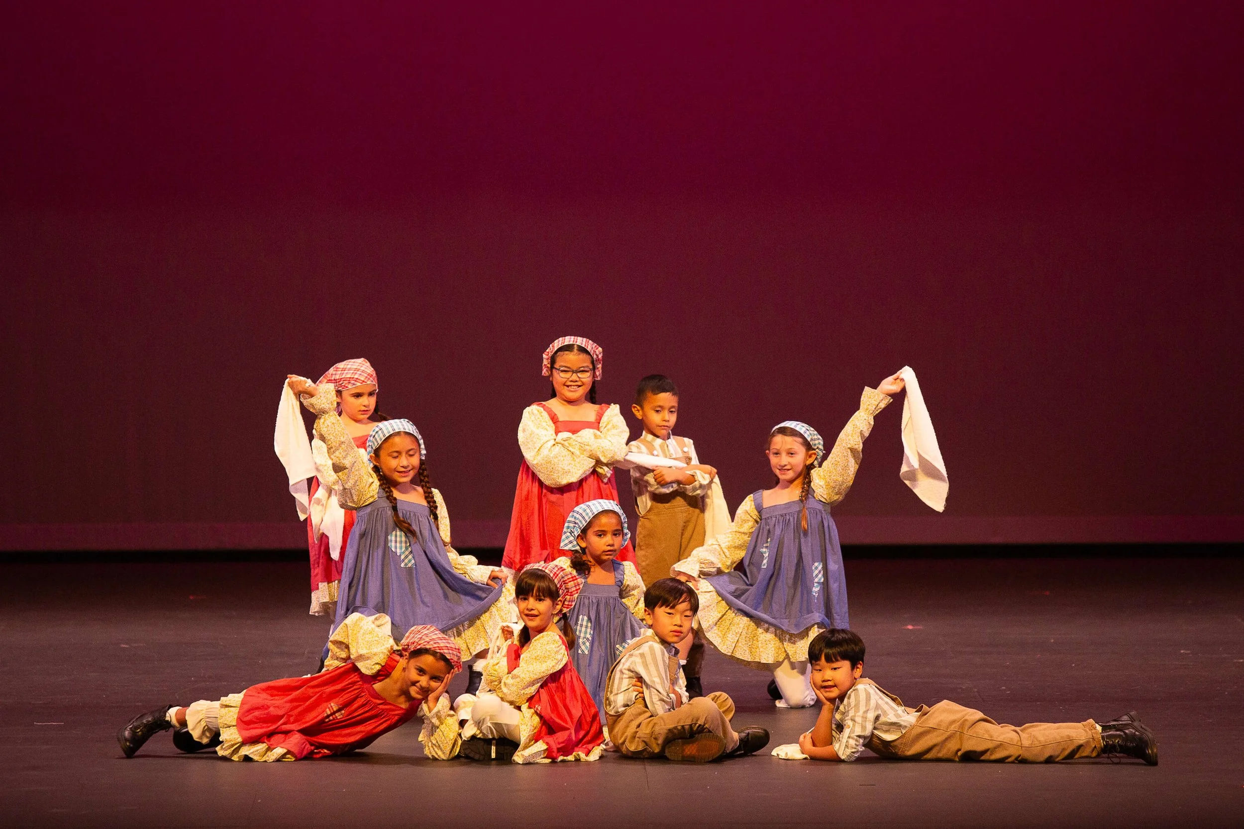 Children in historical costumes performing a dance on stage against a red backdrop.