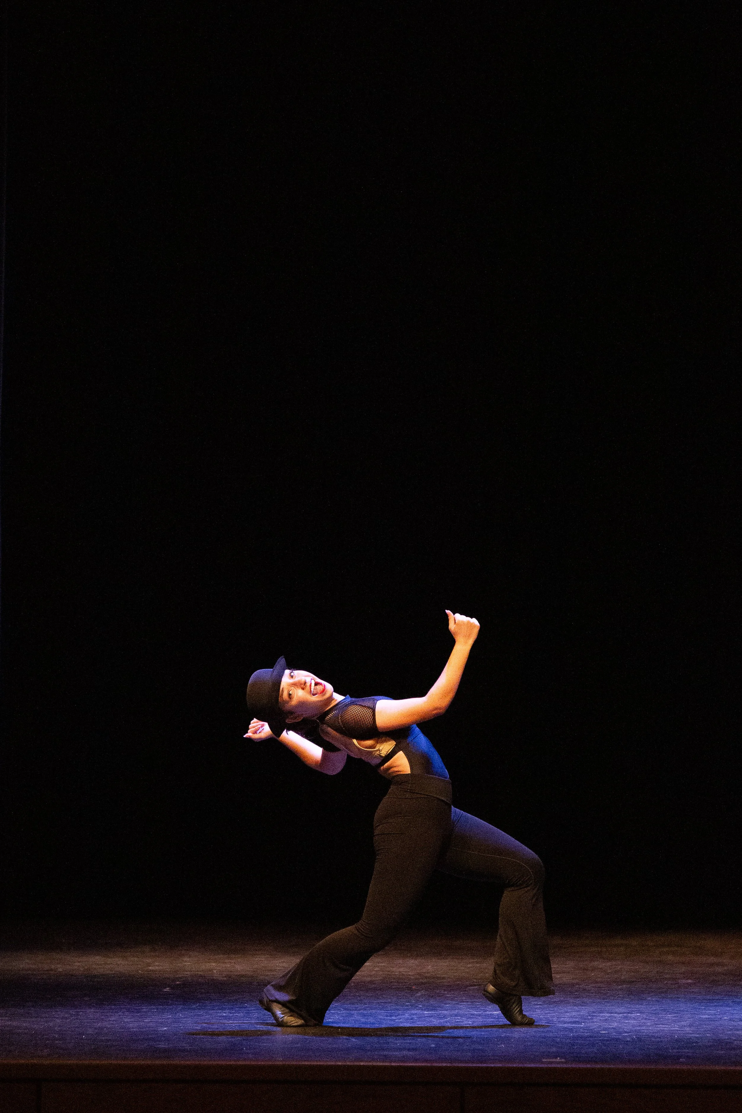 A female dancer in a black hat, black top, and black pants performing on a stage with a dark background. She is in a dance pose with one arm raised and the other bent, smiling at the camera.