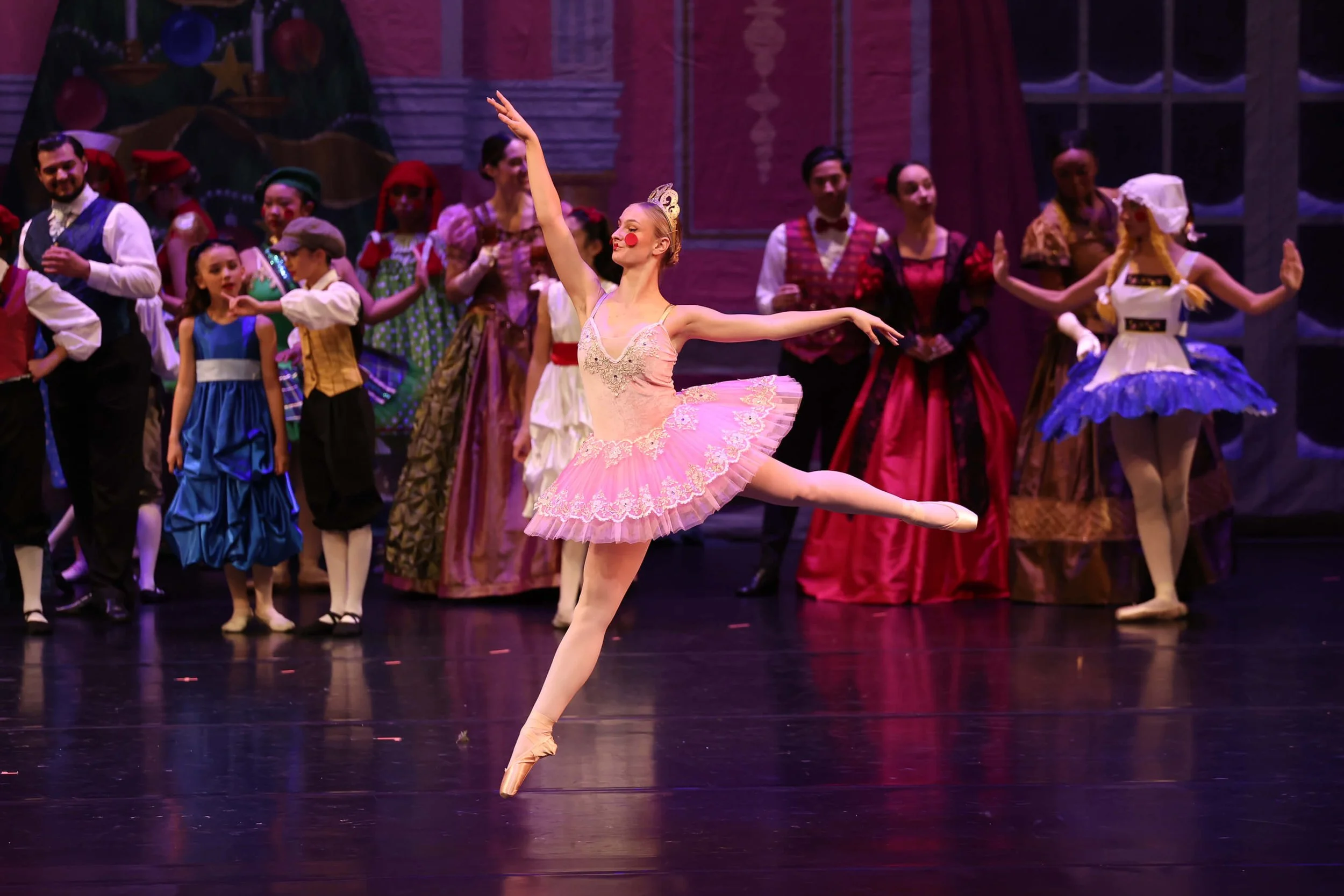 A ballet dancer in a pink tutu performs a arabesque on stage, with a group of cast members in colorful costumes in the background.