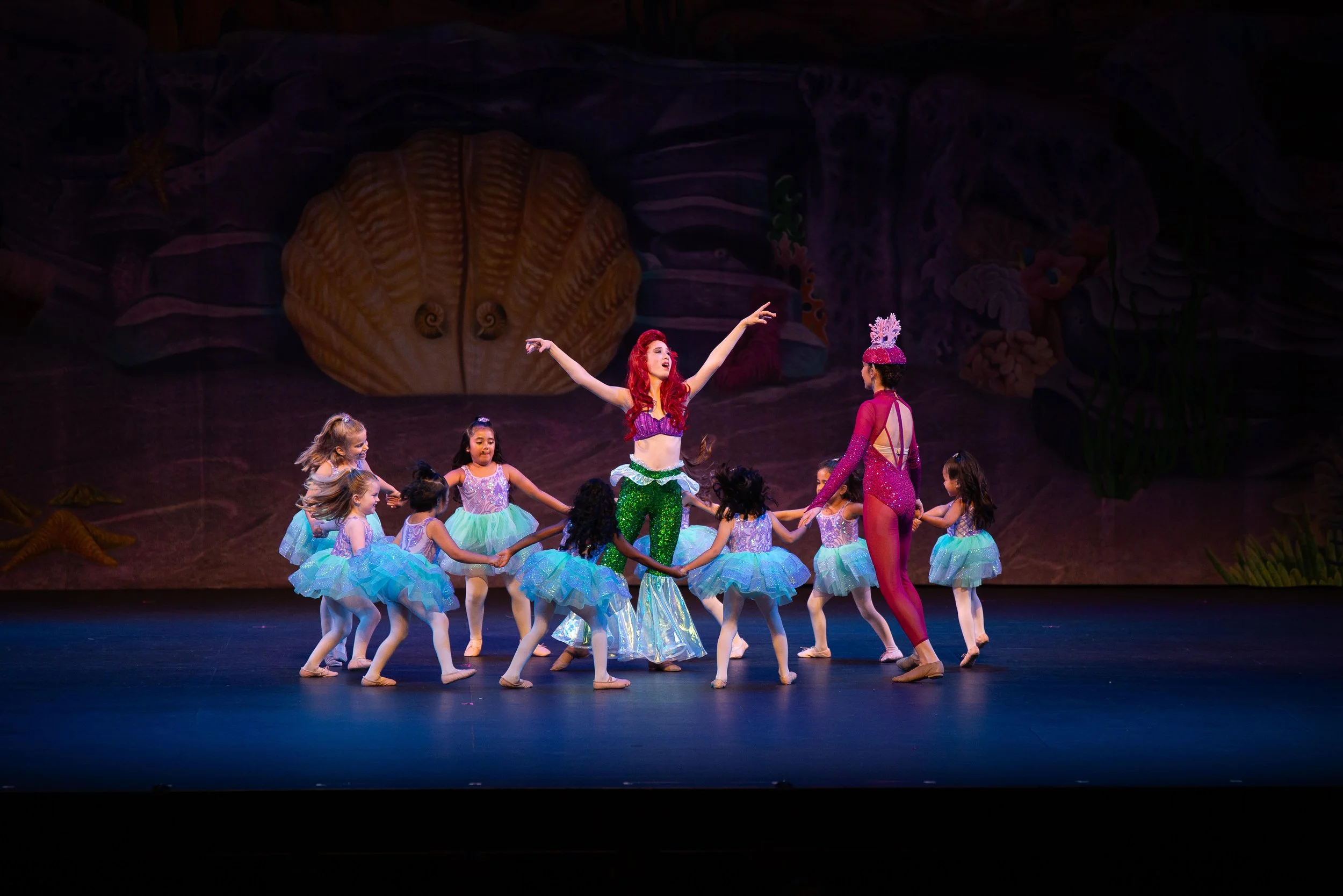 Children's ballet performance on stage featuring dancers in colorful costumes, with a woman dressed as a mermaid in the center, surrounded by children in tutus. The backdrop includes a large seashell and ocean-themed decorations.