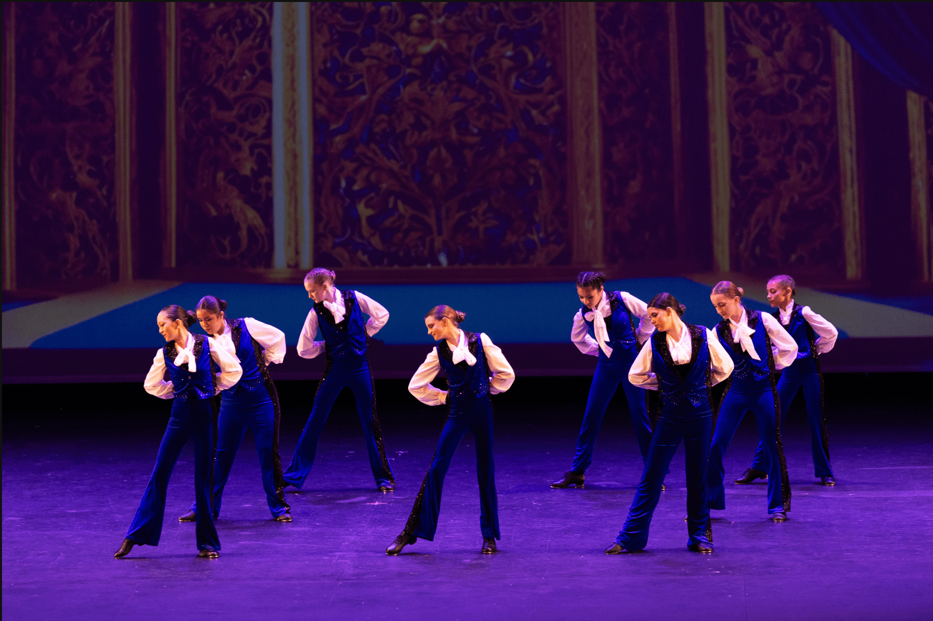 Group of young dancers in blue costumes performing on stage with a decorative background.